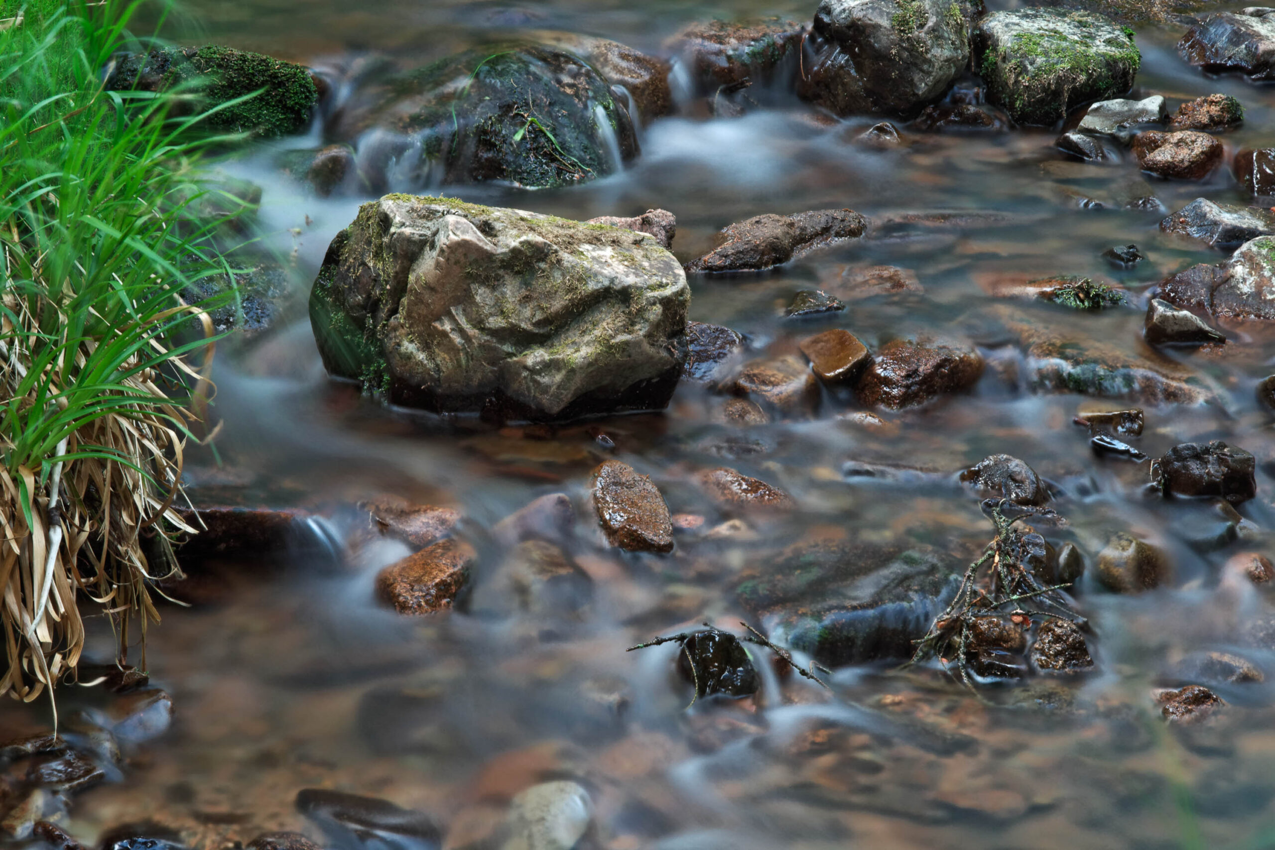 Das Bild zeigt einen fließenden Bach, der über eine steinige Unterlage fließt. Die lange Belichtungszeit erzeugt einen weichen, fließenden Effekt auf dem Wasser. Am Ufer wächst dichtes Grün, das die Szene umrahmt. Die Farben sind gedämpft und wirken beruhigend. Die Szene vermittelt ein Gefühl von Ruhe und Natürlichkeit.