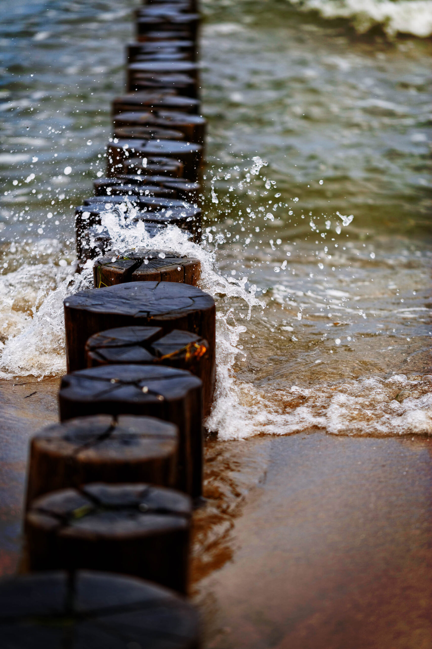 Das Bild zeigt einen Holzstakendeich, der sich in Richtung des Horizonts erstreckt. Das Wasser der Ostsee umspült die Stakendeich-Holzstücke, wobei Gischt und Wellen sichtbar sind. Der Vordergrund besteht aus dunklem Sand, der durch die Bewegung des Wassers leicht verschwommen wirkt. Die Farben sind gedämpft, mit einem Fokus auf Braun-, Grau- und Grüntöne. Das Bild vermittelt eine ruhige und besinnliche Atmosphäre.