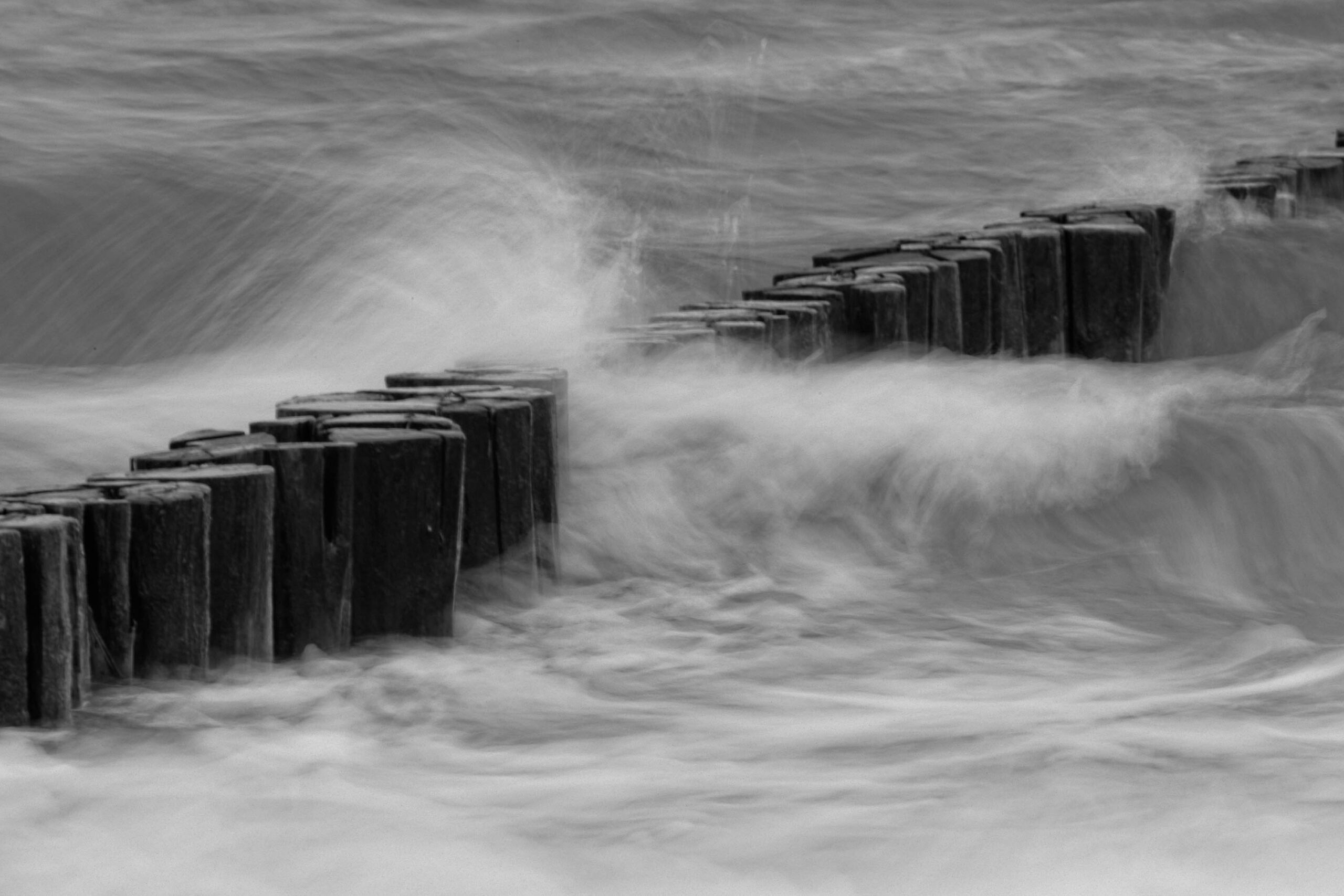 Das vorliegende Schwarzweißfoto zeigt einen hölzernen Wellenbrecher, vermutlich aus Stahlbetonfundamenten mit Holzpfählen, der sich in das stürmische Meer erstreckt. Die lange Belichtungszeit erzeugt einen weichen, fast surrealen Effekt, der die Bewegung des Wassers betont und die Struktur des Wellenbrechers hervorhebt. Die Gischt umspült die Pfähle und erzeugt ein dynamisches Bild. Der Kontext der Aufnahme, Zingst an der Ostsee im Juni 2013, deutet auf eine Küstenlandschaft hin, die für ihre Wellenbrecher bekannt ist. Die Aufnahme fängt die Kraft und Schönheit der Natur ein.