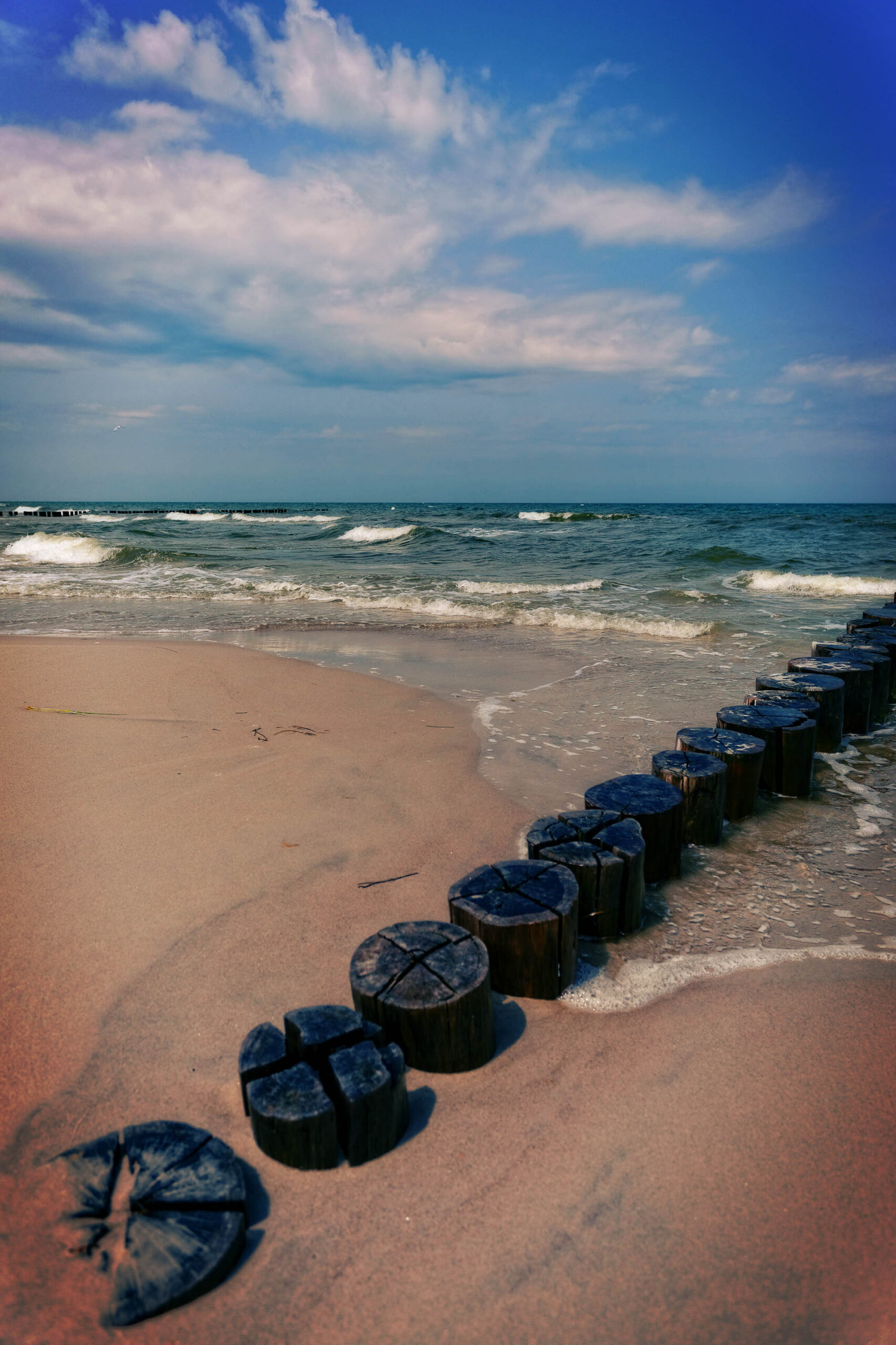 Das Bild zeigt eine Holzsteg-Anlage, die sich durch das flache Wasser eines Sandstrandes erstreckt. Die Stege bestehen aus dicken, abgetretenen Baumstämmen, die in regelmäßigen Abständen angeordnet sind. Der Strand ist sandig und hell, während das Wasser in verschiedenen Blautönen schimmert. Der Himmel ist bewölkt, mit vereinzelten hellen Wolken. Die Szene vermittelt eine ruhige und friedliche Atmosphäre.
