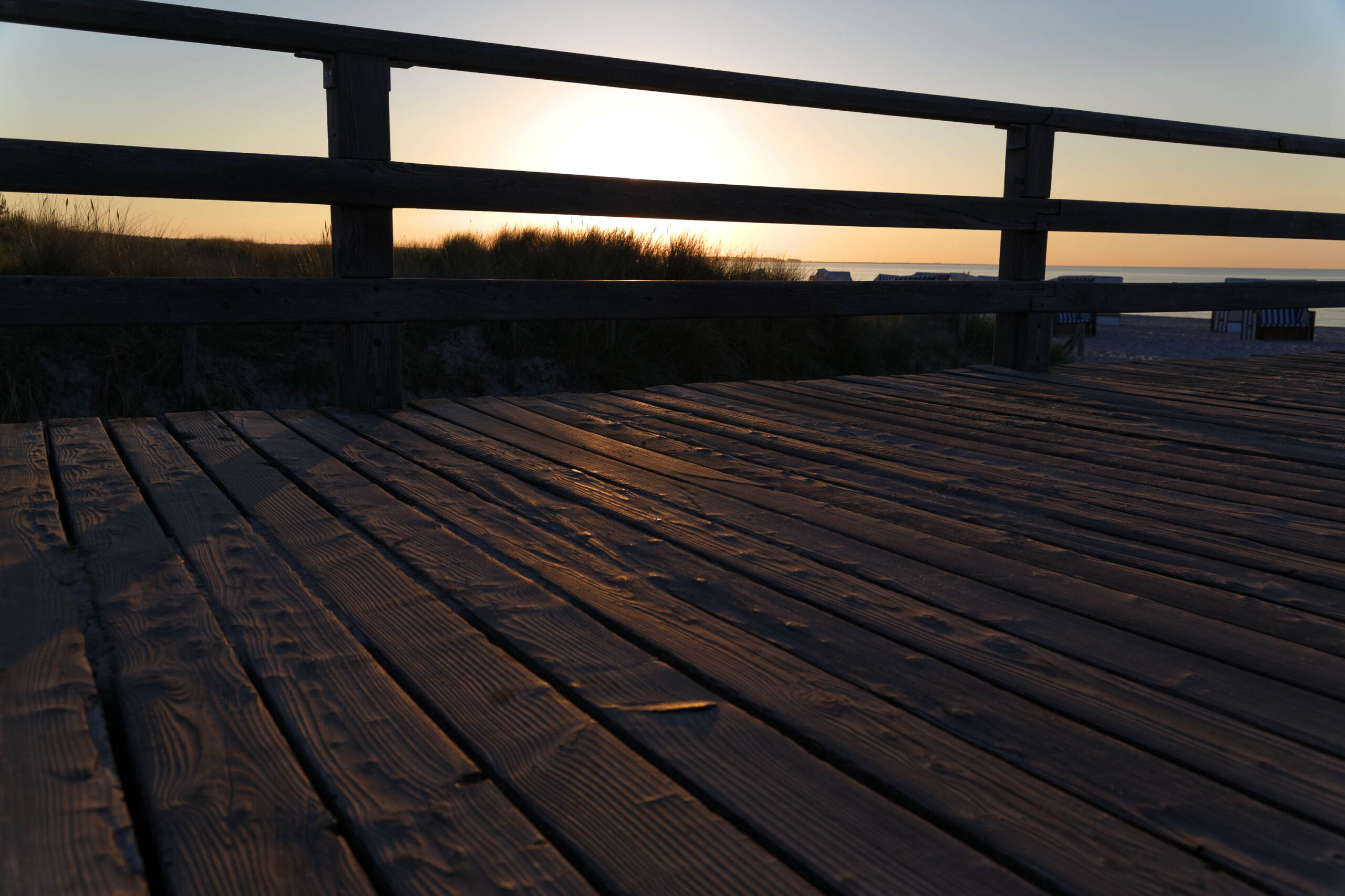 Das Bild zeigt eine Nahaufnahme eines Holzdecks, vermutlich Teil einer Promenade oder eines Weges durch ein Naturschutzgebiet. Die Holzplanken sind dunkel und zeigen eine deutliche Maserung. Das Licht des Sonnenuntergangs fällt schräg auf das Deck und erzeugt Reflexionen und Schatten, die die Textur des Holzes hervorheben. Im Hintergrund sind Dünen und ein paar Gebäude erkennbar, die den Horizont bilden. Der Himmel ist in warmen Farben gehalten, was auf einen Sonnenuntergang hindeutet. Die Szene vermittelt eine ruhige und friedliche Atmosphäre.
