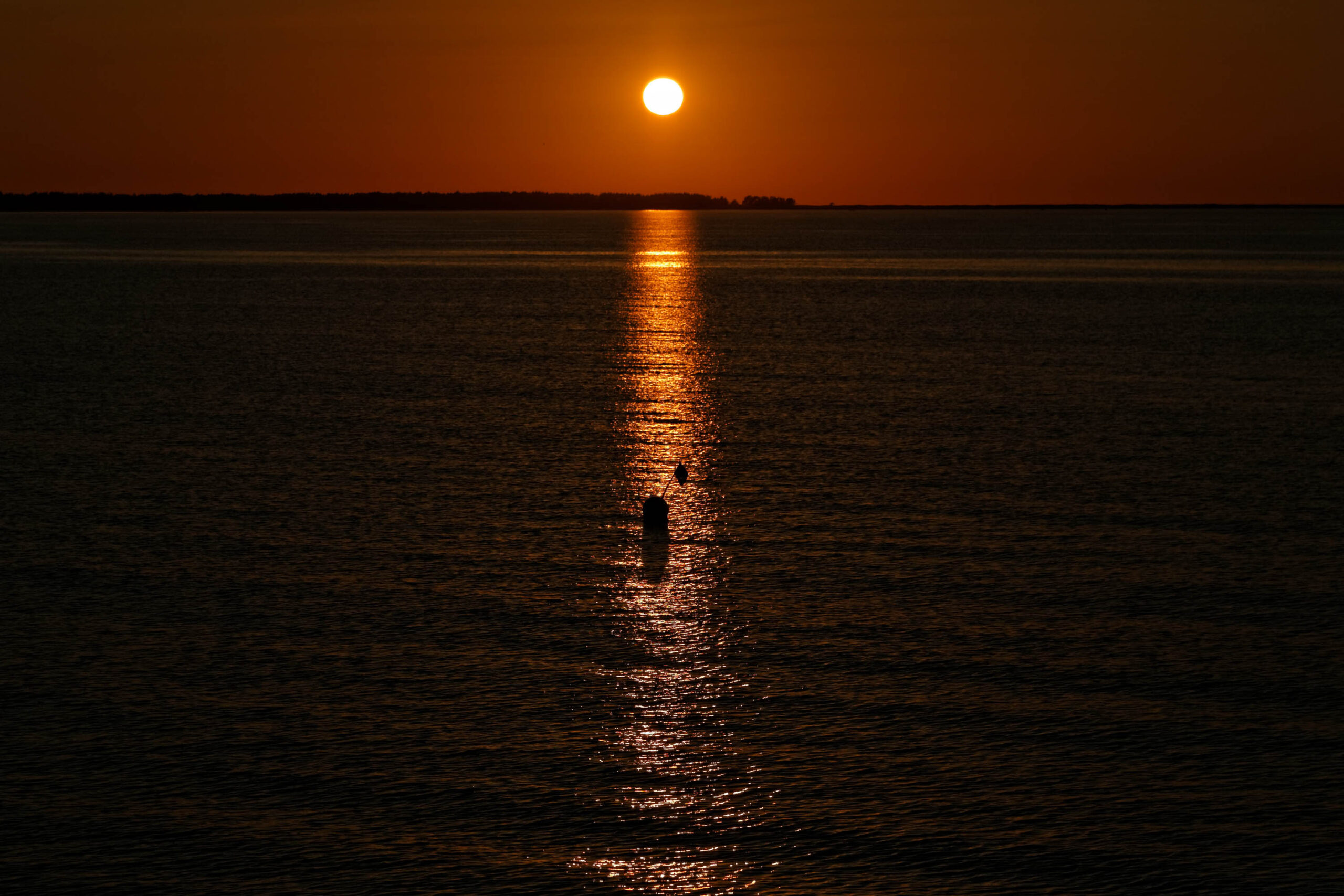 Das Bild zeigt einen Sonnenuntergang über dem Wasser, wahrscheinlich der Ostsee, aufgenommen im Juni 2013 in der Nähe von Prerow, Darß. Die Sonne steht tief am Horizont und erzeugt einen hellen, goldenen Lichtstreifen auf der Wasseroberfläche. Drei Personen sind im Vordergrund zu sehen, silhouettiert gegen das Licht. Der Himmel ist in warmen Orange- und Rottönen gehalten. Die Landschaft am Horizont ist nur schemenhaft erkennbar.