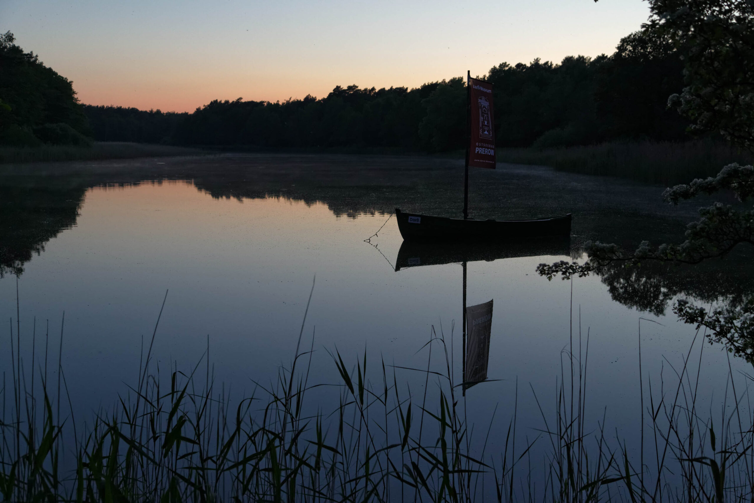 Die Aufnahme zeigt einen ruhigen See bei Sonnenuntergang. Im Mittelpunkt steht ein Windrad, dessen Silhouette sich deutlich im Wasser spiegelt. Die Umgebung besteht aus dichtem, dunklem Wald, der sich am Horizont abzeichnet. Im Vordergrund ragen Schilfhalme auf, die die Szene strukturieren. Der Himmel ist in sanften Farben des Sonnenuntergangs gehalten, was eine ruhige und friedliche Atmosphäre erzeugt. Die Spiegelung im Wasser verstärkt den Eindruck von Ruhe und Symmetrie.