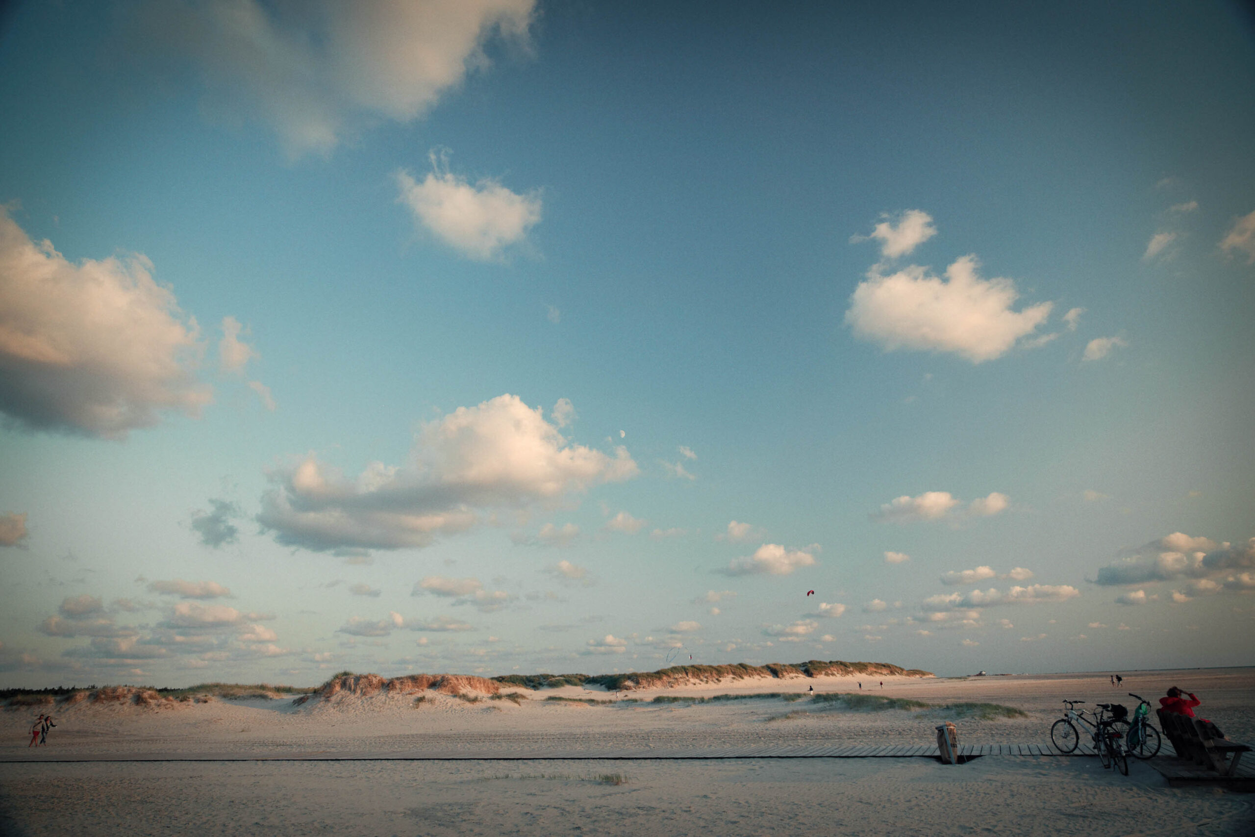 Die Aufnahme zeigt eine typische Szene von St. Peter-Ording, einem Seebad in Schleswig-Holstein. Der Sandstrand erstreckt sich weitläufig, und im Hintergrund ragen die charakteristischen Dünen auf. Der Himmel ist blau und von weißen Wolken durchzogen, was die Szene hell und freundlich wirken lässt. Mehrere Personen sind auf dem Strand zu sehen, einige entspannen sich, andere scheinen sich sportlichen Aktivitäten zu widmen. Eine Gruppe von Fahrrädern steht am Rand des Strandes, was auf die Beliebtheit des Radfahrens in der Region hindeutet. Die Aufnahme fängt die entspannte und friedliche Atmosphäre dieses beliebten Urlaubsziels ein.