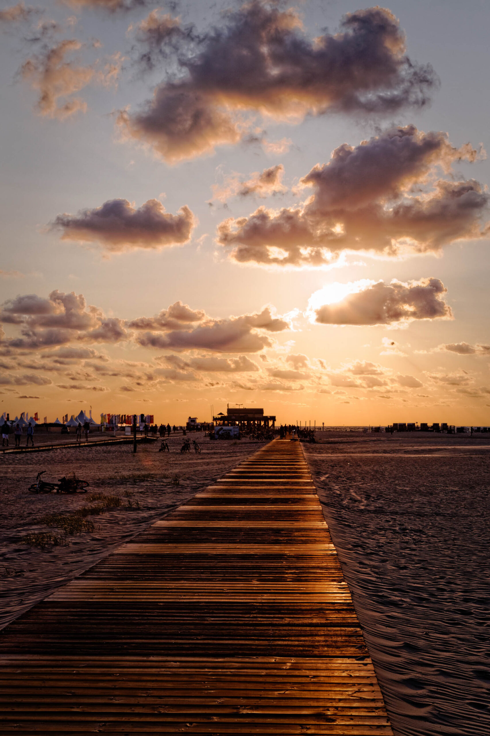 Das Foto zeigt einen hölzernen Steg, der sich in die Ferne erstreckt und in den Strand von St. Peter-Ording führt. Der Himmel ist von einem dramatischen Sonnenuntergang mit tiefroten und orangefarbenen Tönen sowie zahlreichen Wolken bedeckt. Im Hintergrund sind Menschen und Strandaufbauten erkennbar. Die Szene vermittelt eine ruhige und friedliche Atmosphäre.
