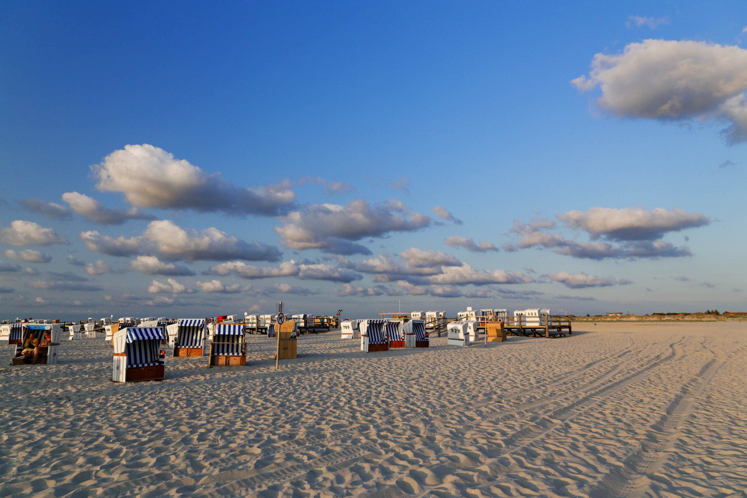 Das Bild zeigt eine weite Ansicht eines Sandstrandes in St. Peter-Ording. Eine lange Reihe von typischen, gestreiften Strandkörbe erstreckt sich über den Strand. Der Sand ist hell und weist deutliche Spuren von Wellen und Fußabdrücken auf. Der Himmel ist strahlend blau mit vereinzelten, weißen Wolken. Die Szene vermittelt eine friedliche und entspannte Atmosphäre. Die Perspektive ist von unten aufgenommen, was die Weite des Strandes betont.