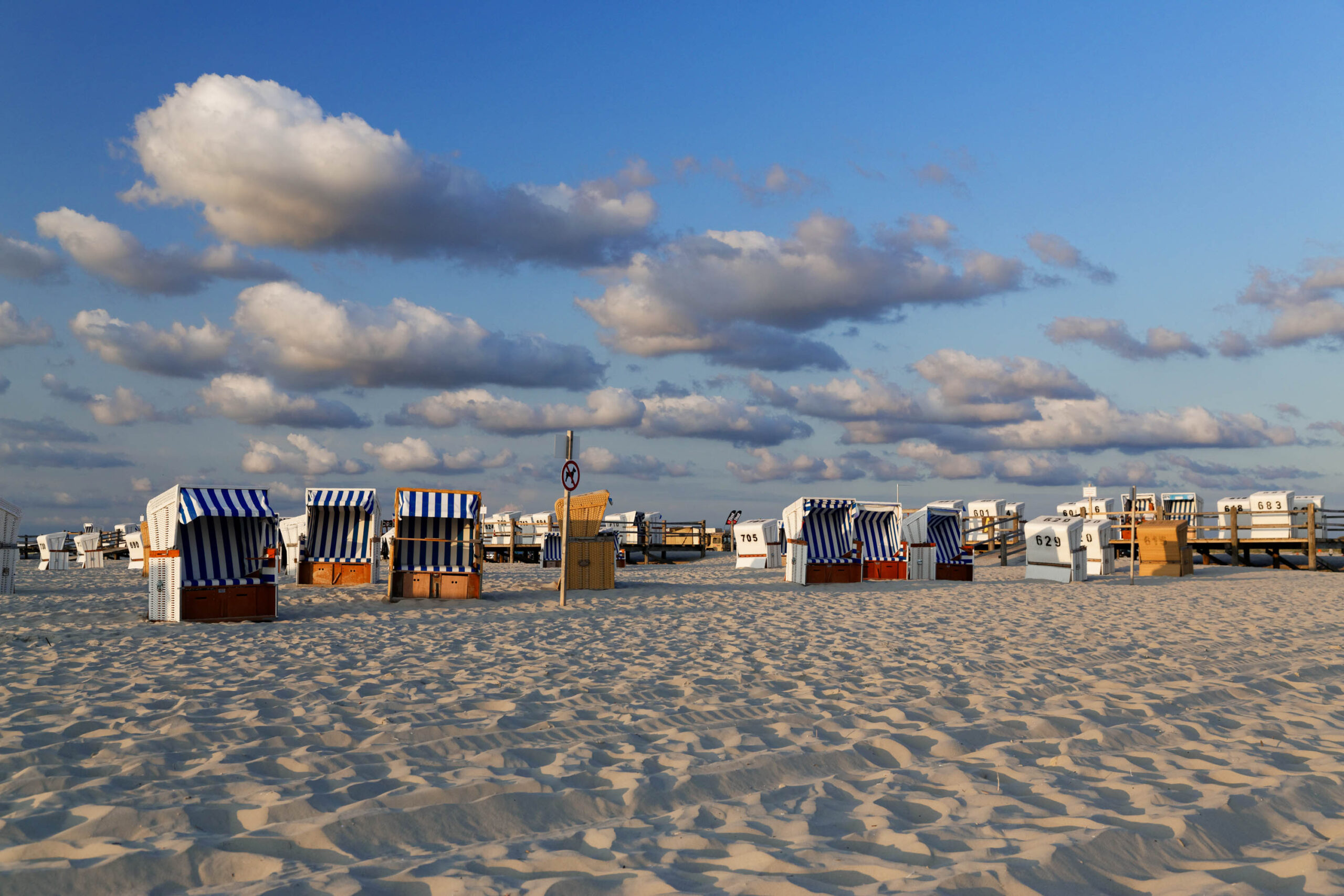 Das Bild zeigt eine typische Szene von St. Peter-Ording, einer norddeutschen Seebadestadt. Eine Reihe von farbenfrohen Strandkörben steht dicht an dicht auf dem hellen Sandstrand. Die Strandkörbe sind in verschiedenen Farben gestreift und tragen Nummern, was auf die Vergabe der Strandplätze hindeutet. Der Himmel ist blau und von weißen Wolken bedeckt, was auf ein leicht bewölkten Tag schließen lässt. Der Sand ist fein und hell, und die Szene vermittelt ein Gefühl von Ruhe und Erholung.