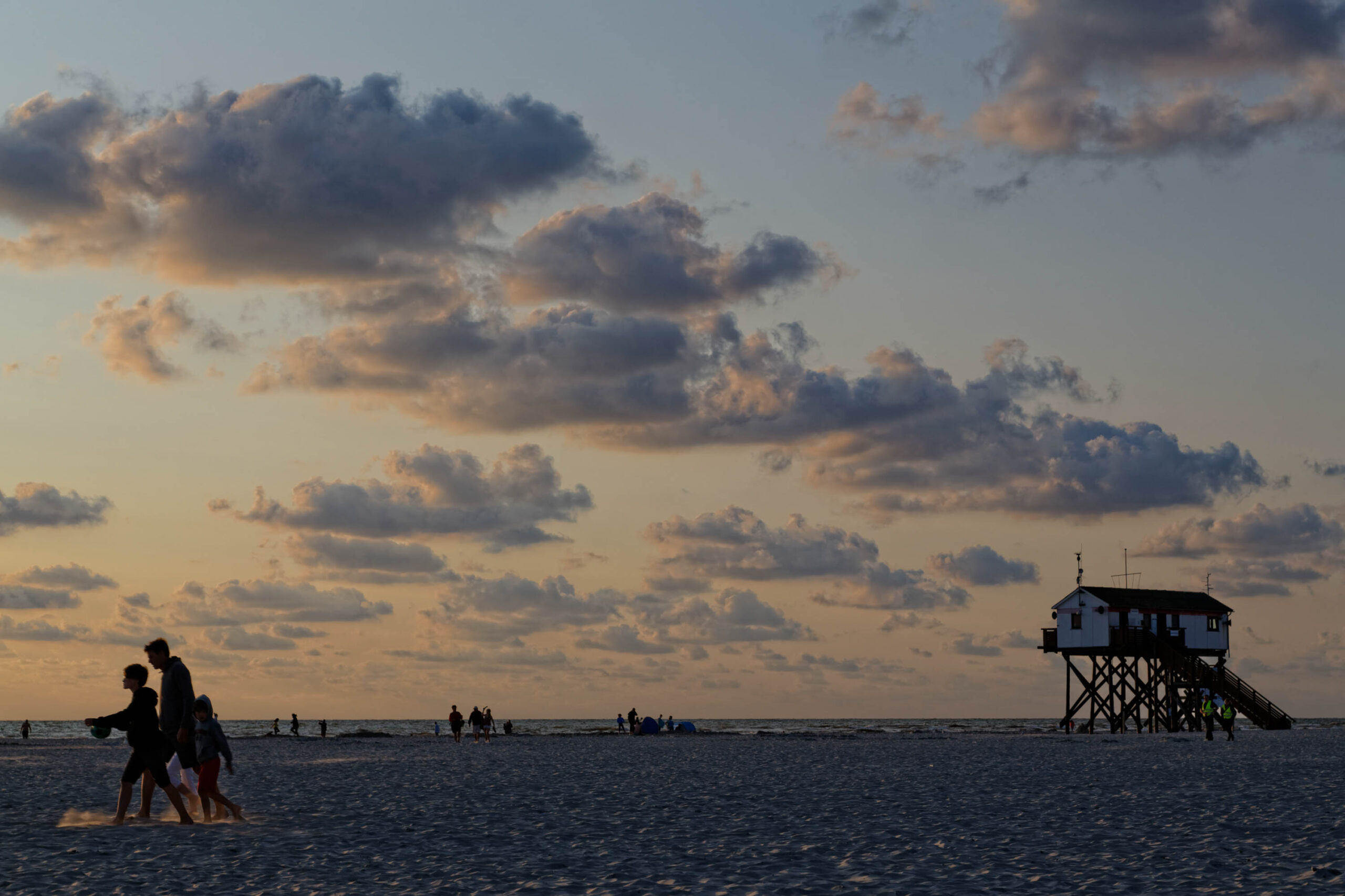 Die Aufnahme zeigt einen Pfahlbau, der sich im Abendlicht am Strand von St. Peter-Ording erhebt. Der Himmel ist von einem warmen Farbton durchzogen, der sich in den ruhigen Wasseroberfläche widerspiegelt. Im Vordergrund sind mehrere Personen zu sehen, die am Strand entlanggehen, wobei eine Familie im Fokus steht. Die Szene vermittelt eine friedliche und entspannte Atmosphäre, typisch für einen Sommerabend am Nordsee-Strand.