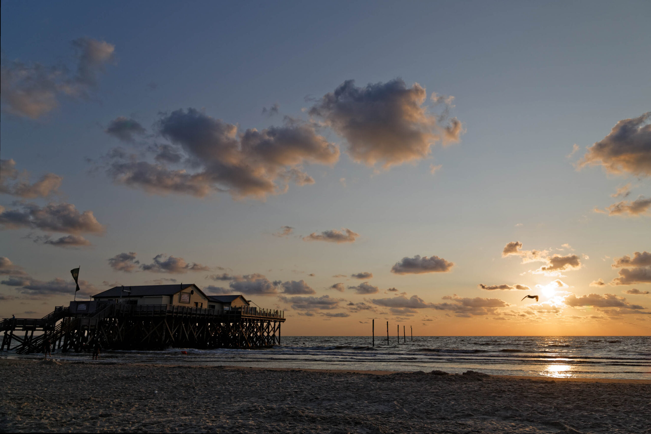 Die Aufnahme zeigt einen Pfahlbau, der sich auf Stelzen über den Sandstrand erhebt. Der Himmel ist in warmen Farben von Orange und Gelb gefärbt, was auf einen Sonnenuntergang hindeutet. Einige vereinzelte Wolken ziehen über den Himmel. Das Wasser ist ruhig und spiegelt die Farben des Himmels wider. Im Vordergrund ist der Sandstrand zu sehen, der von vereinzelten Grashalmen bedeckt ist. Einige Vögel fliegen über dem Wasser. Die Szene vermittelt eine friedliche und entspannte Atmosphäre.