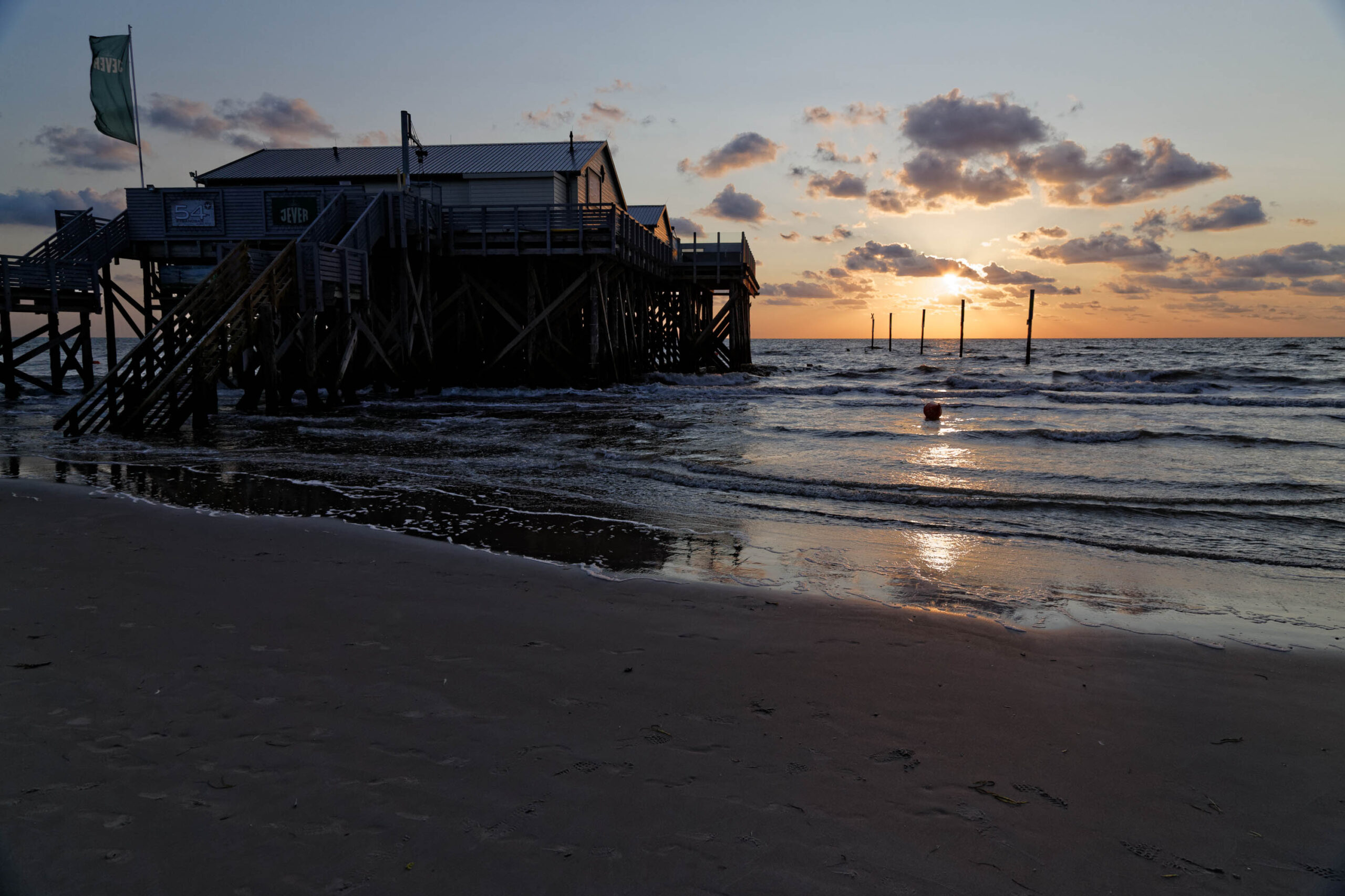Das Bild zeigt einen Pfahlbau, der auf Stelzen in der Nordsee steht. Der Himmel ist von einem warmen Sonnenuntergang in Orange- und Rosatönen gefärbt, der sich im Wasser spiegelt. Der Strand ist dunkel und sandig. Im Vordergrund befindet sich eine Person, die im Wasser steht. Die Szene vermittelt eine friedliche und entspannte Atmosphäre.