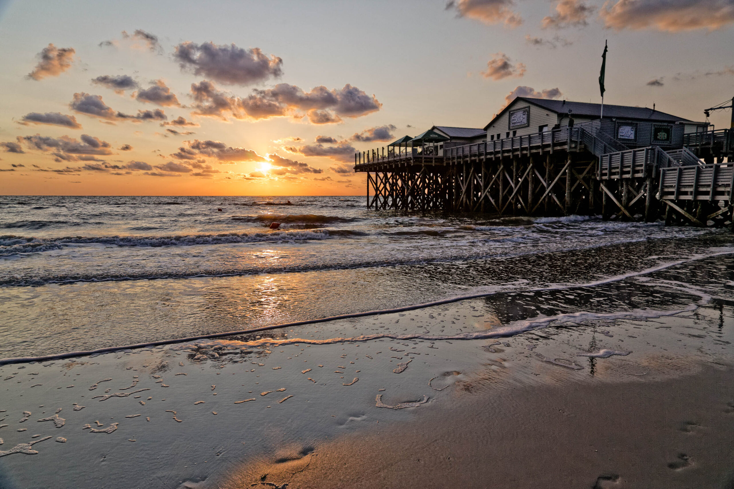 Das Bild zeigt einen Pfahlbau, der auf Stelzen in das Meer gebaut wurde. Der Himmel ist von einem warmen Sonnenuntergang in Orange- und Rosatönen gefärbt. Der Strand ist feucht und reflektiert das Licht. Das Bild vermittelt eine friedliche und entspannte Atmosphäre.