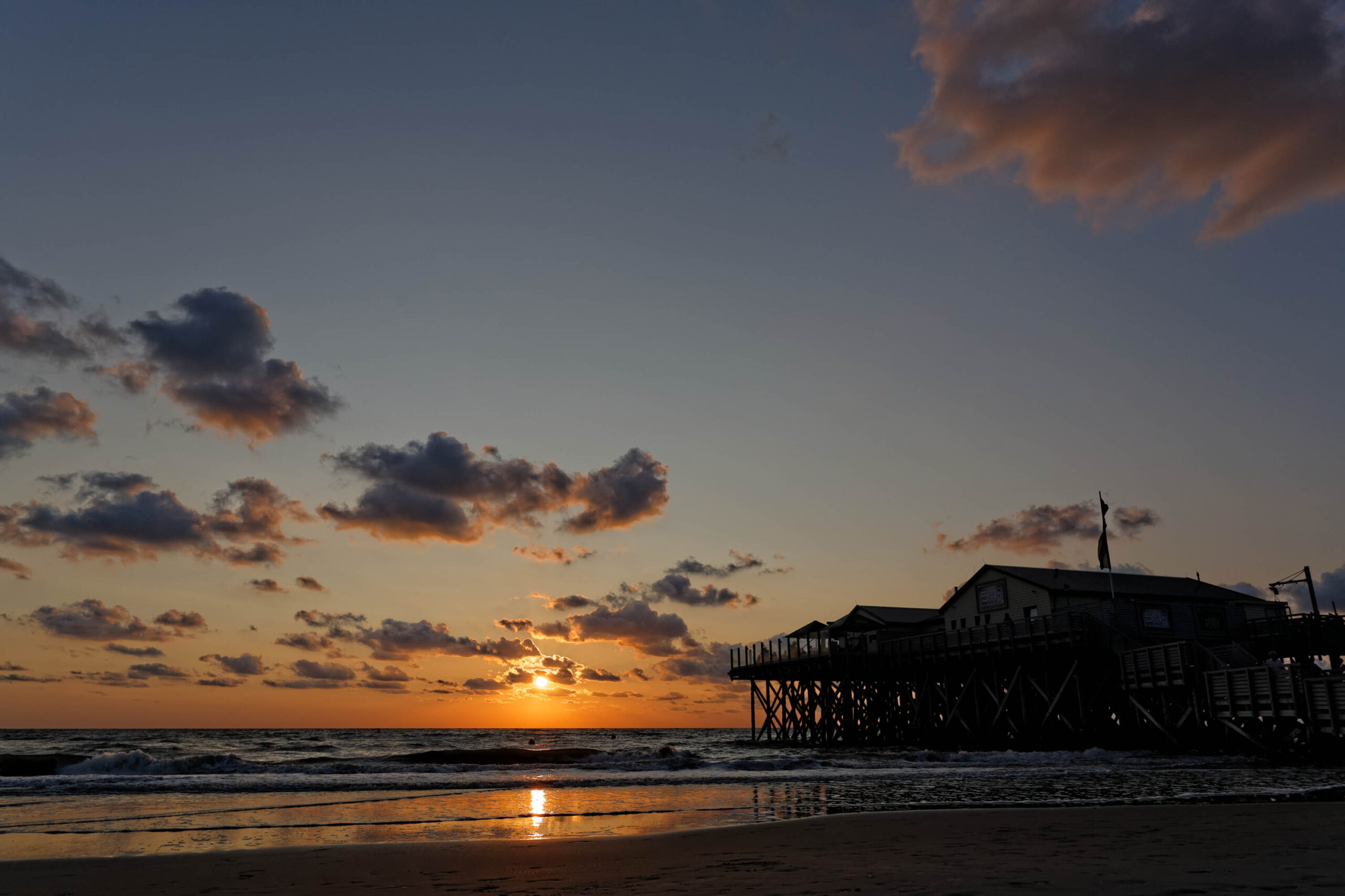 Das Bild zeigt einen Pfahlbau am Strand von St. Peter-Ording bei Sonnenuntergang. Der Himmel ist bedeckt mit Wolken, die in warmen Farben leuchten. Das Wasser ist ruhig und spiegelt das Licht der untergehenden Sonne wider. Der Pfahlbau ist im Vordergrund zu sehen und steht auf Stelzen im Wasser. Der Strand ist dunkel und sandig. Die Szene vermittelt eine ruhige und friedliche Atmosphäre.