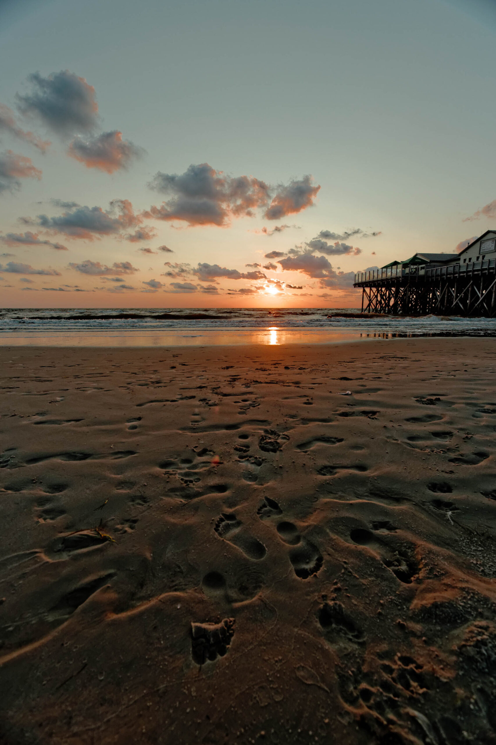 Das Bild zeigt einen Pfahlbau auf dem Ordinger Strand in St. Peter-Ording bei Sonnenuntergang. Der Strand ist mit Fußabdrücken übersät, die im feuchten Sand sichtbar sind. Der Himmel ist in warmen Farben von Orange und Rosa gefärbt, wobei die untergehende Sonne durch Wolken hindurchscheint. Der Pfahlbau steht auf Stelzen im Wasser und scheint in das Licht der untergehenden Sonne getaucht zu sein. Die Szene vermittelt eine friedliche und entspannte Atmosphäre.