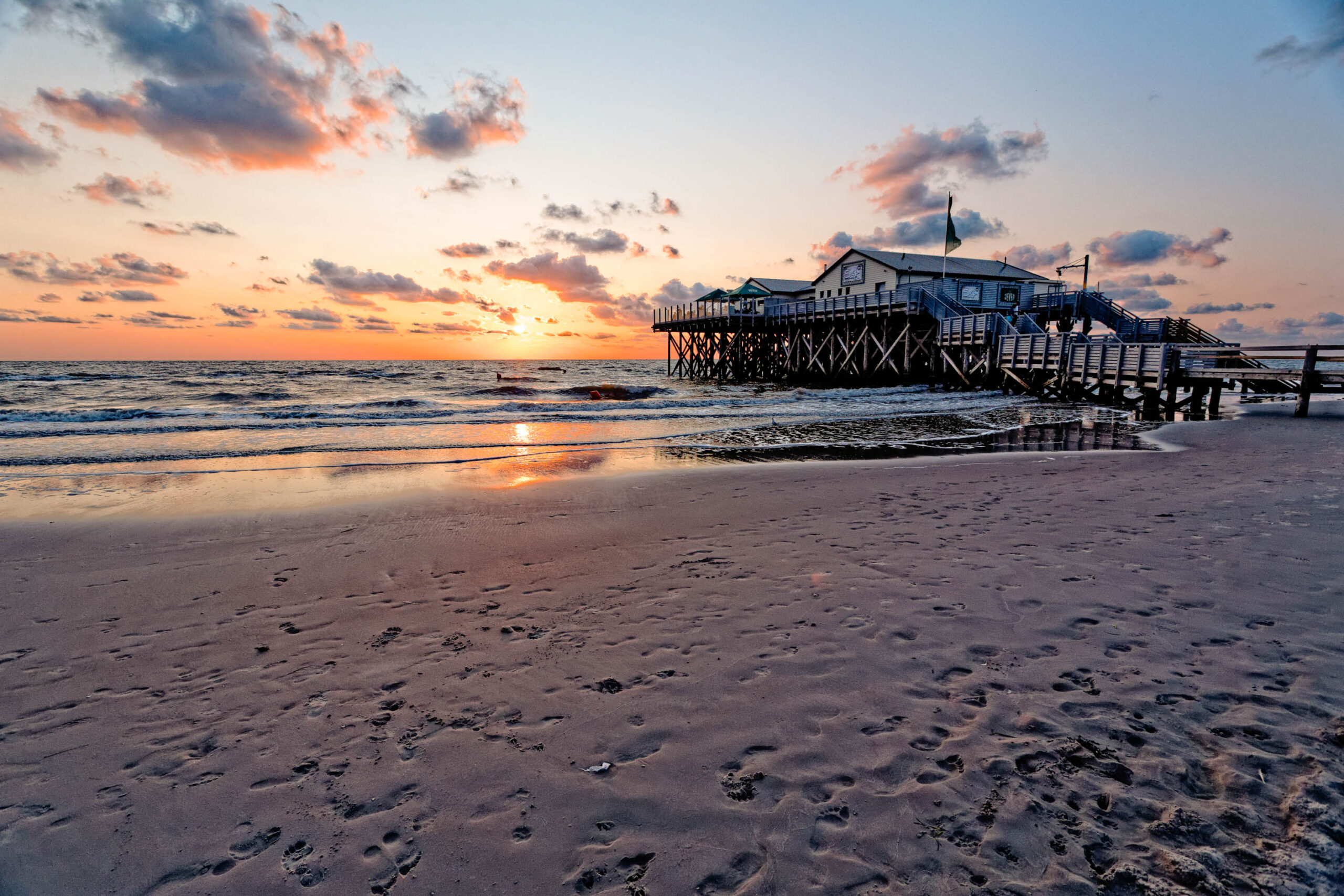 Die Aufnahme zeigt einen Pfahlbau, der sich auf dem Strand von St. Peter-Ording erhebt. Der Bau steht auf langen Pfählen, die ihn über den Sand und das Wasser heben. Der Himmel ist in warmen Farben von Orange, Rosa und Violett gefärbt, was auf einen Sonnenuntergang hindeutet. Der Strand ist mit feuchtem Sand bedeckt, der die Farben des Himmels reflektiert. Das Licht der untergehenden Sonne erzeugt eine stimmungsvolle Atmosphäre. Die Aufnahme wurde wahrscheinlich im Sommer aufgenommen, da die Bedingungen mild und angenehm erscheinen.