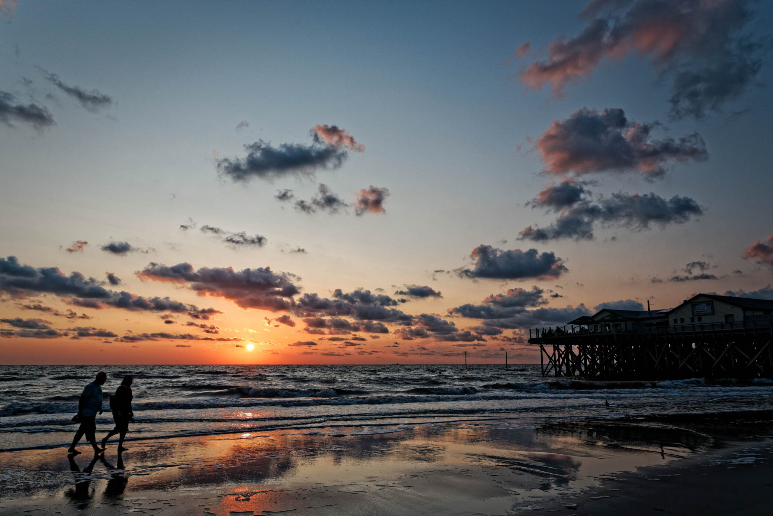Das Bild zeigt ein romantisches Szenario am Strand von St. Peter-Ording bei Sonnenuntergang. Ein Paar ist im Vordergrund zu sehen, wie es Hand in Hand am Strand entlangspaziert. Der Strand ist feucht und reflektiert das orangefarbene Licht der untergehenden Sonne. Im Hintergrund erhebt sich ein Pfahlbau, der typisch für die Küste von St. Peter-Ording ist. Der Himmel ist bedeckt mit Wolken, die das Licht der Sonne in dramatischen Farben erscheinen lassen. Die Szene vermittelt eine friedliche und entspannte Atmosphäre.