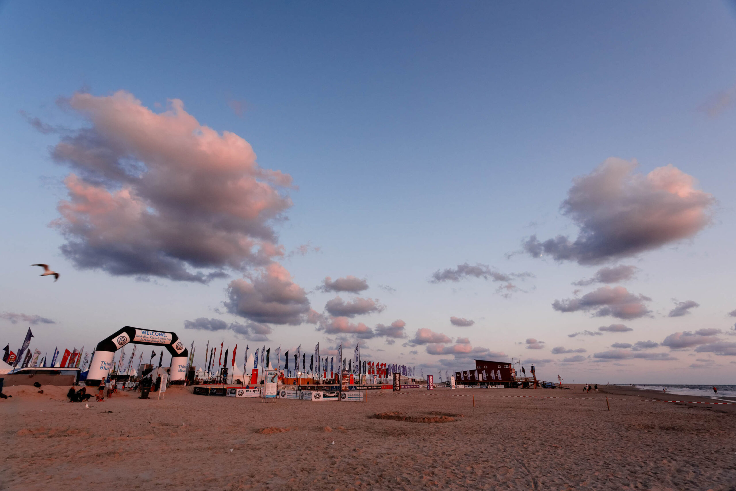 Die Aufnahme zeigt einen weiten Blick auf den Ordinger Strand in St. Peter-Ording bei Sonnenuntergang. Im Vordergrund erstreckt sich der feuchte, dunkle Sandstrand. Im mittleren Bereich des Bildes reihen sich zahlreiche Pfahlbauten aneinander, die mit bunten Flaggen geschmückt sind. Diese Pfahlbauten sind typisch für den Ordinger Strand und beherbergen Restaurants, Cafés und Geschäfte. Im Hintergrund ist der helle Horizont mit dem Meer zu sehen. Der Himmel ist von einem tiefen Blau mit vereinzelten, weißen Wolken bedeckt, die das Licht der untergehenden Sonne reflektieren. Einige Möwen fliegen über den Strand. Die Szene vermittelt eine friedliche und entspannte Atmosphäre.