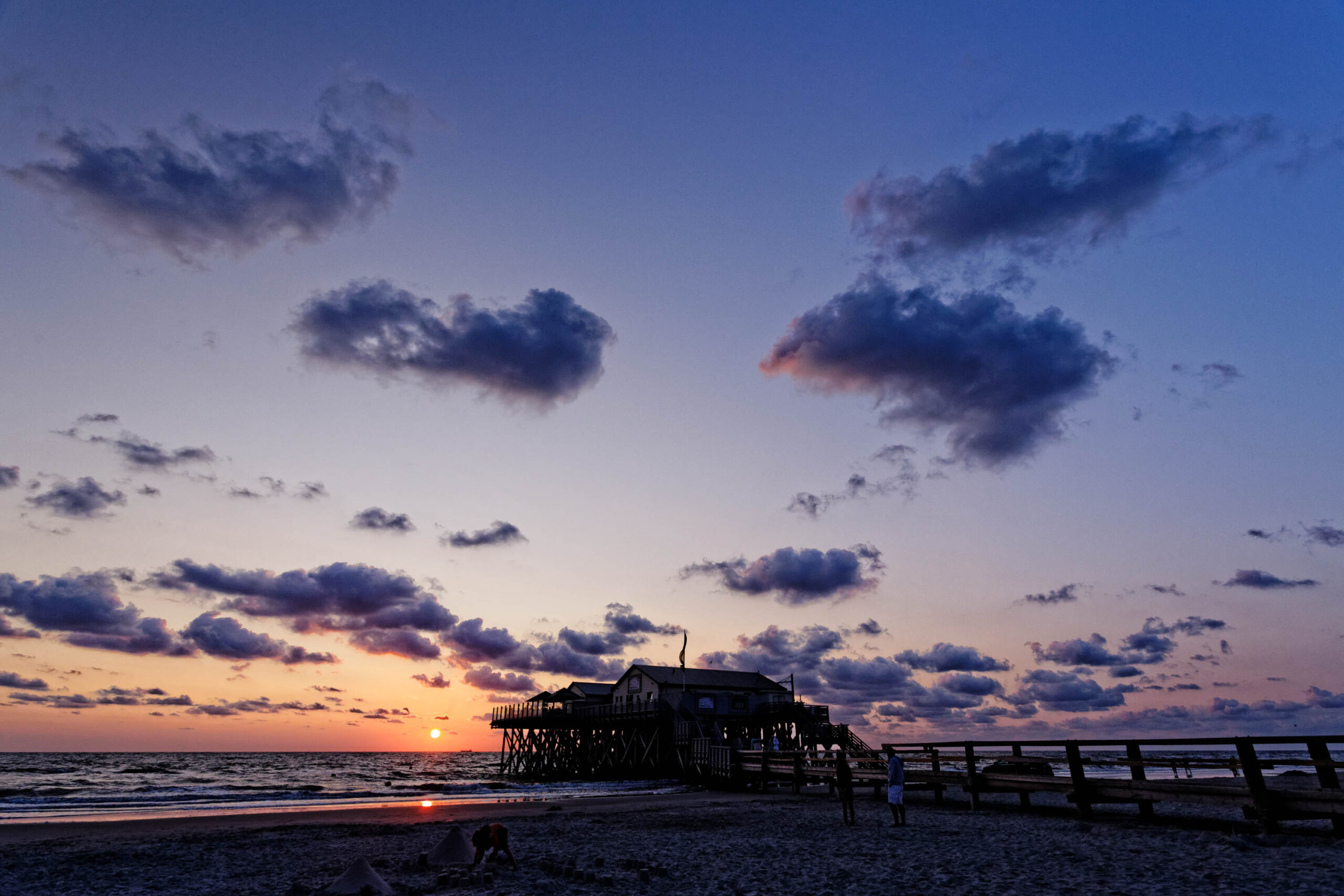 Ein atemberaubendes Foto fängt einen Pfahlbau bei Sonnenuntergang am Strand von St. Peter-Ording ein. Der Himmel ist in einem Farbenspiel aus Lila, Rosa und Orange gehalten, während die Sonne langsam im Meer versinkt. Der Pfahlbau, ein charakteristisches Merkmal der Küste, steht silhouettiert vor dem Himmel. Einige Personen sind auf dem Strand zu sehen, die den Sonnenuntergang genießen. Der Strand ist mit feinem Sand bedeckt, der im warmen Licht glänzt. Die Szene vermittelt eine friedliche und entspannte Atmosphäre.