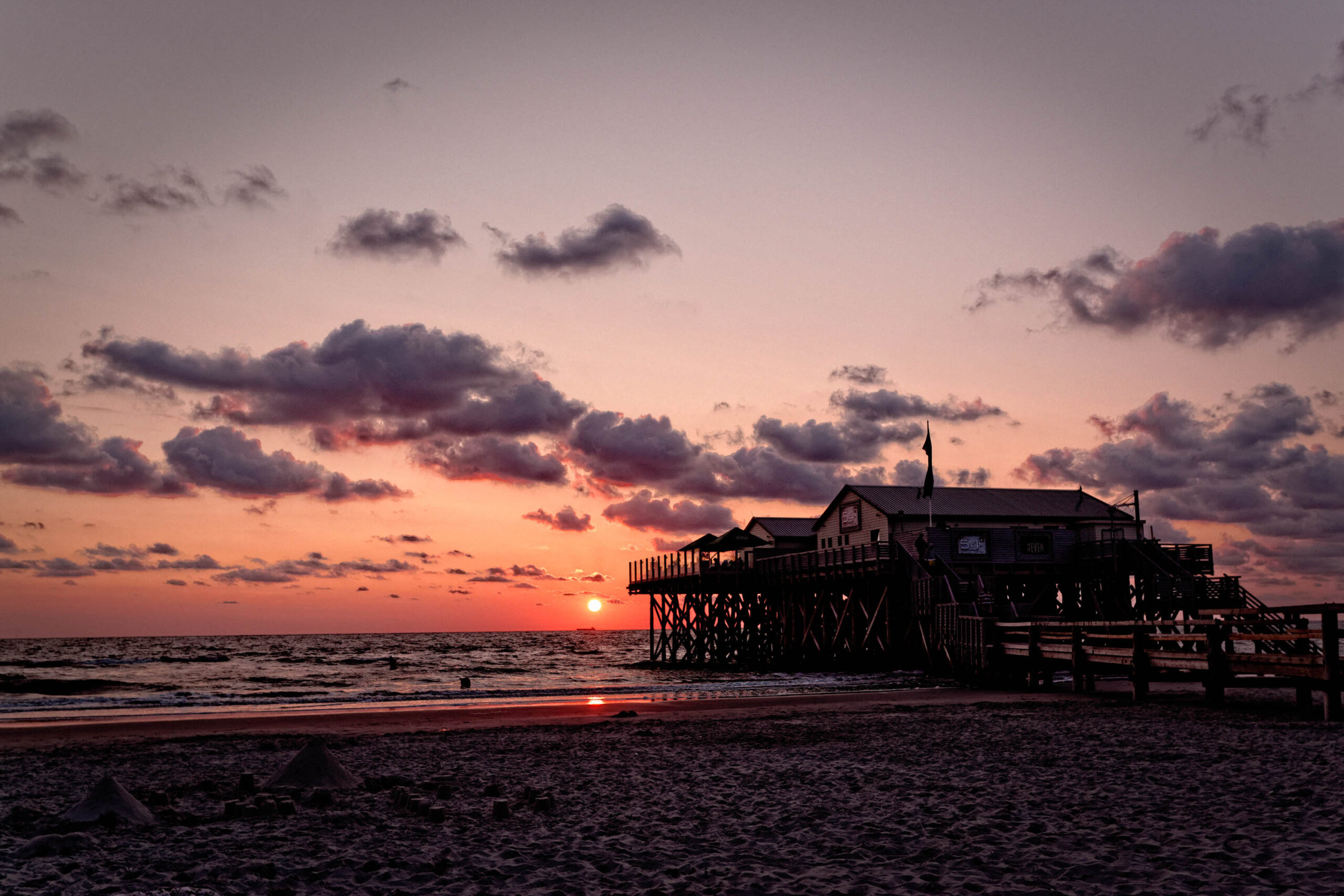 Das Bild zeigt einen Pfahlbau, der sich auf dem Ordinger Strand in St. Peter-Ording erhebt. Der Himmel ist in dramatischen Rottönen und Violetttönen gefärbt, was auf einen Sonnenuntergang hindeutet. Das Wasser der Nordsee ist ruhig und spiegelt die Farben des Himmels wider. Der Strand ist mit dunklem Sand bedeckt, der mit Treibholz übersät ist. Der Pfahlbau ist im Vordergrund und dominiert die Szene. Er ist im Dunkeln gehalten, was die Aufmerksamkeit auf den Himmel lenkt. Die Szene vermittelt eine friedliche und ruhige Atmosphäre.