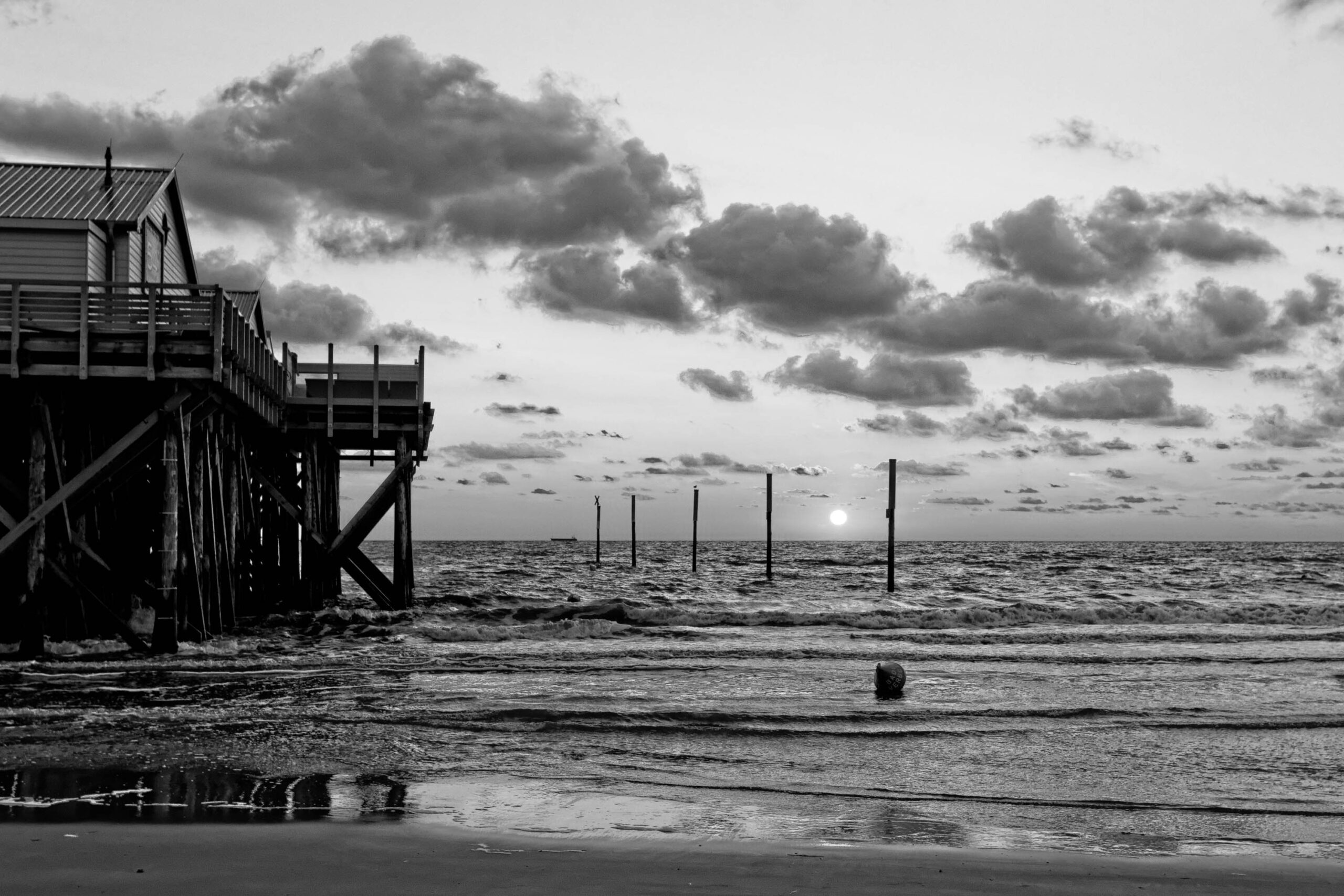 Das Foto zeigt einen Pfahlbau, der sich über das Wasser erstreckt, mit einem dunklen Himmel und einem Sonnenuntergang im Hintergrund. Der Pfahlbau ist aus Holz und hat mehrere Plattformen und Treppen. Das Wasser ist ruhig und reflektiert den Himmel. Der Strand ist sandig und leer. Die Szene ist ruhig und friedlich.