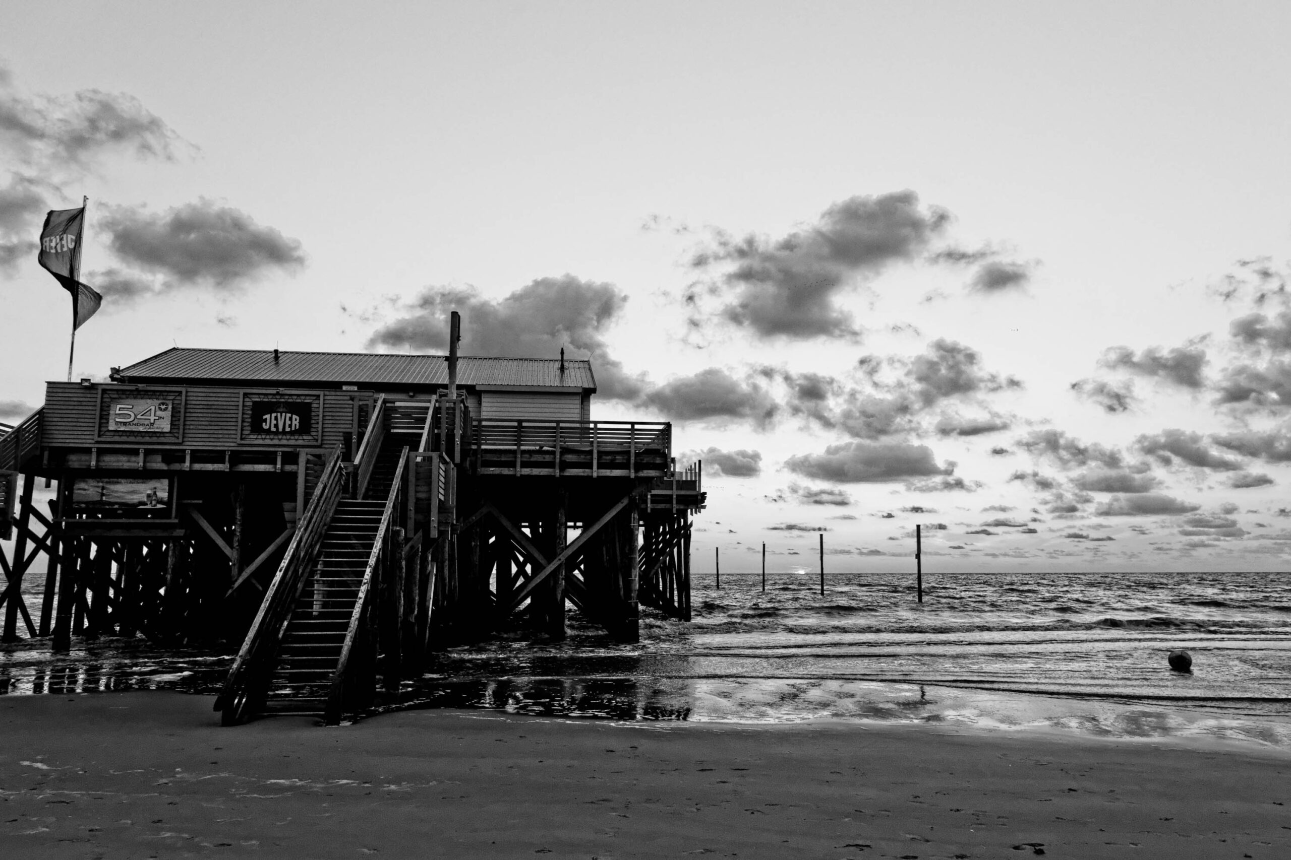 Das Foto zeigt einen Pfahlbau, der auf Stelzen über den Strand ragt. Der Bau ist aus Holz und hat eine einfache Struktur mit einem Satteldach. Eine Treppe führt vom Strand zum Eingang des Gebäudes. Im Hintergrund ist das Meer zu sehen, das in dunklen Wellen an den Strand rollt. Der Himmel ist bewölkt und erzeugt eine düstere Atmosphäre. Ein einzelner Schwimmer ist im Wasser zu sehen. Die Szene vermittelt ein Gefühl von Ruhe und Abgeschiedenheit.