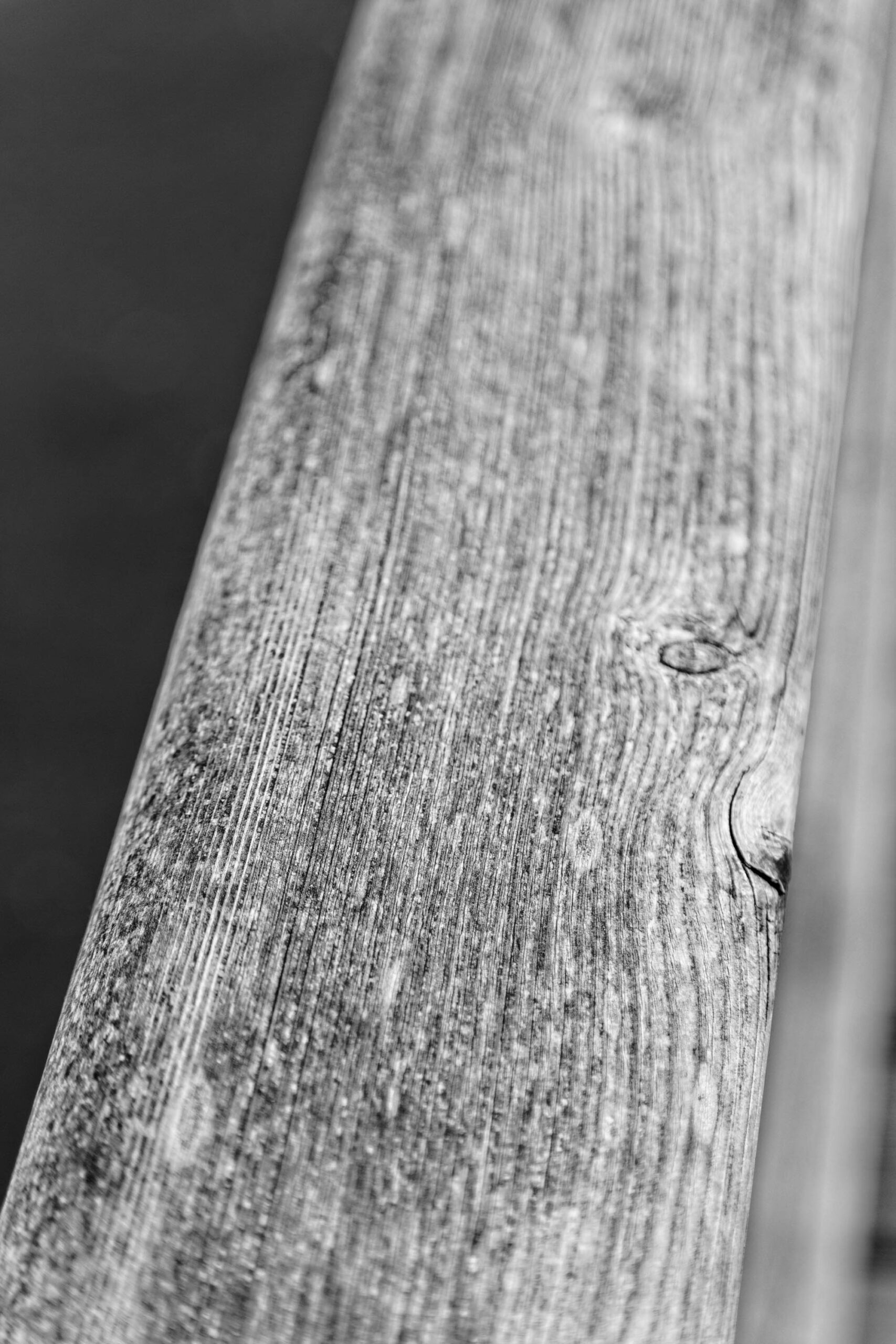 Das Bild zeigt eine detailreiche Nahaufnahme eines Holzpfostens. Die Maserung des Holzes ist deutlich sichtbar, ebenso wie Risse und Unebenheiten, die auf seine Alterung und Witterungseinflüsse hindeuten. Der Pfosten ist schräg gestellt und wird von einem dunklen Hintergrund abgesetzt, was die Textur und die Details des Holzes hervorhebt. Die Schwarz-Weiß-Darstellung verstärkt den Kontrast und betont die raue Beschaffenheit des Materials. Der Pfosten scheint Teil einer größeren Struktur zu sein, die im Bild nicht vollständig zu erkennen ist. Die Komposition konzentriert sich auf die Details der Holzoberfläche und erzeugt eine minimalistische und abstrakte Ästhetik.