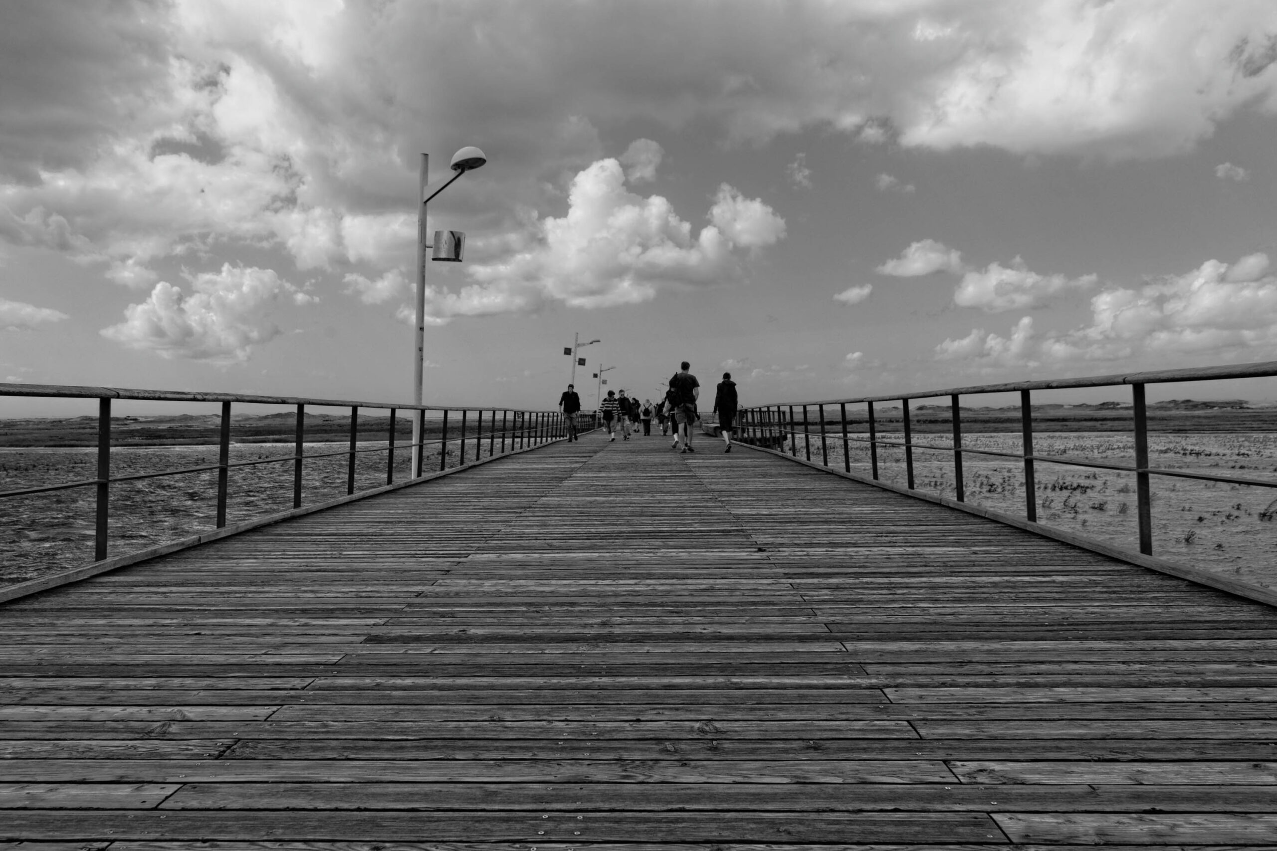Die Fotografie zeigt eine Perspektive entlang eines Holzstegs in St. Peter-Ording, Nordsee. Der Steg erstreckt sich in gerader Linie in die Ferne, wobei die Perspektive die Länge des Stegs betont. Mehrere Personen sind auf dem Steg zu sehen, die sich in Richtung des Horizonts bewegen. Der Himmel ist von dichten Wolken bedeckt, was eine dramatische Atmosphäre schafft. Die Schwarz-Weiß-Darstellung verstärkt die Textur des Holzes und die Kontraste im Bild.