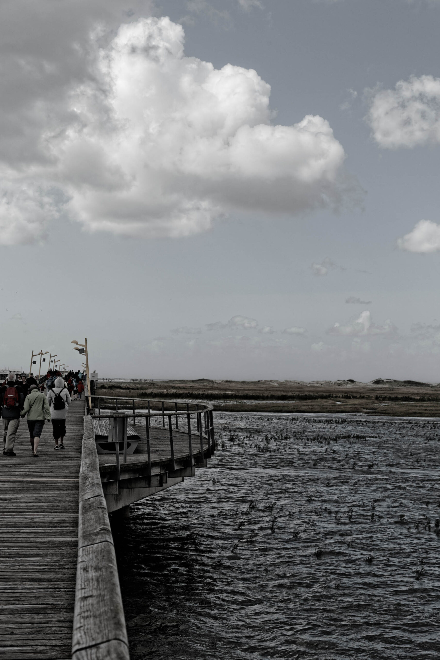 Das vorliegende Schwarz-Weiß-Foto zeigt einen Ausschnitt einer Seebrücke in St. Peter-Ording, an der Nordsee. Die Brücke führt über ein ruhiges, dunkles Gewässer, das vermutlich ein Küstenbereich oder ein flaches Meer ist. Im Hintergrund erstreckt sich eine Dünenlandschaft, die durch die monochrome Darstellung eine gewisse Dramatik erhält. Mehrere Personen sind auf der Brücke zu sehen, die in Richtung des Horizonts spazieren. Der Himmel ist bewölkt, was die Szene in ein melancholisches Licht taucht. Die Komposition betont die Weite der Landschaft und die Isolation der Menschen in der Natur.