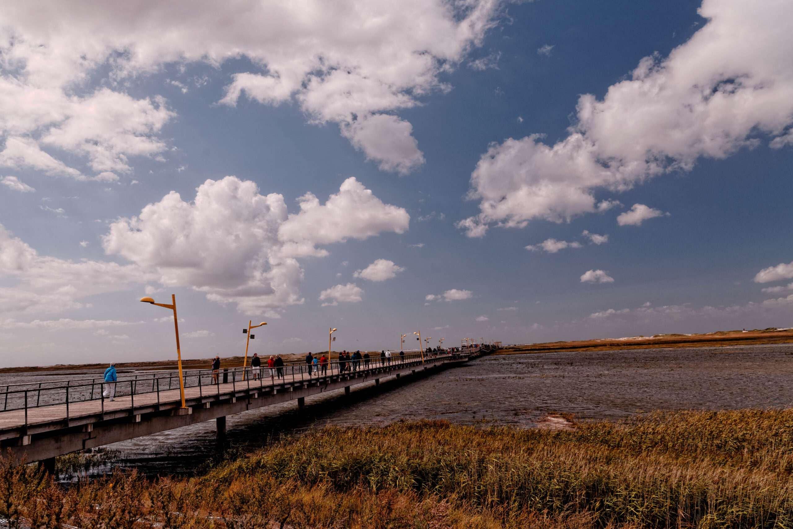 Das Bild zeigt eine lange Seebrücke in St. Peter-Ording, Nordsee. Die Brücke erstreckt sich weit in eine ruhige Bucht, die von trockenem, braunem Gras gesäumt ist. Der Himmel ist blau und von weißen, bauschigen Wolken bedeckt. Mehrere Personen befinden sich auf der Brücke und entlang des Ufers. Die Szene vermittelt eine friedliche und entspannte Atmosphäre.
