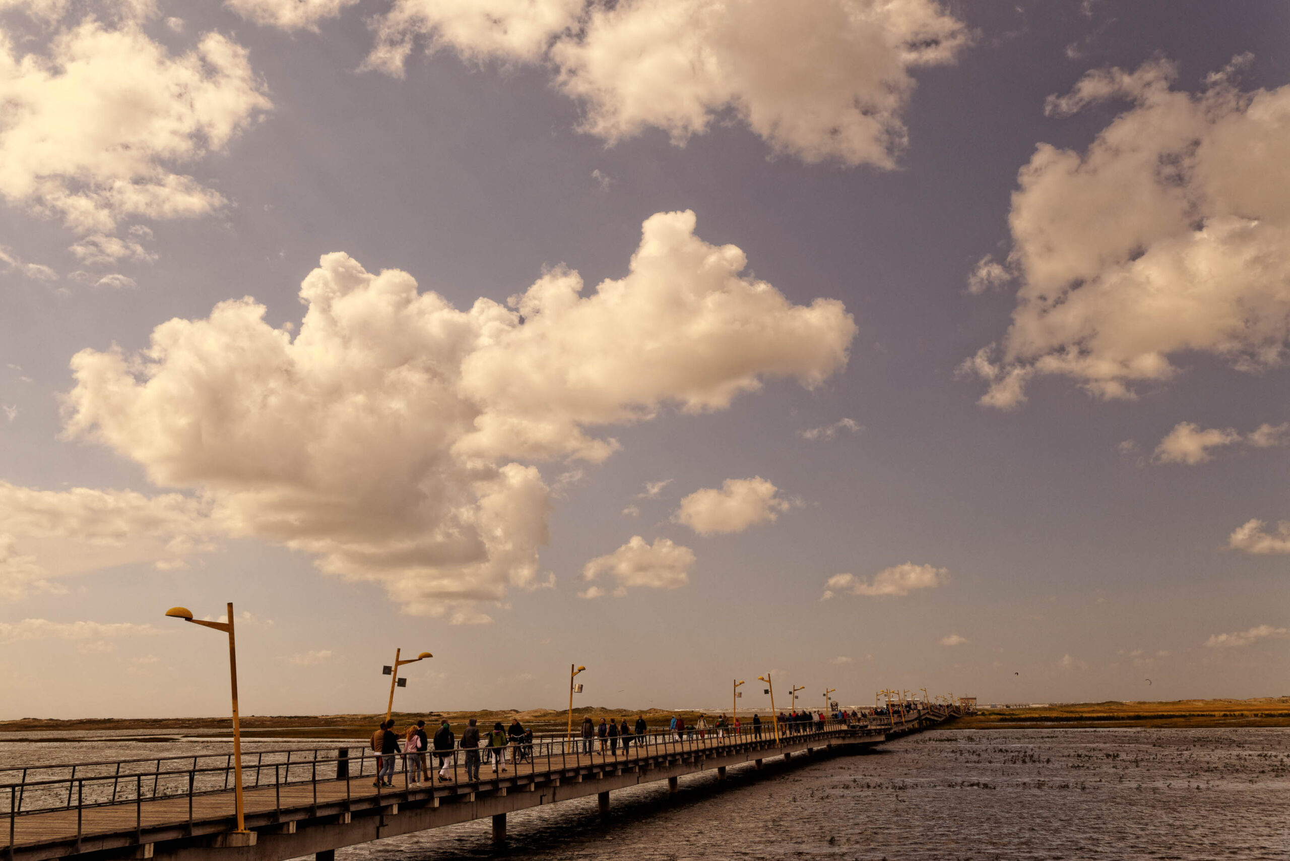 Das Foto zeigt einen Blick auf die Seebrücke in St. Peter-Ording, Nordsee. Die Brücke erstreckt sich weit ins Meer hinein und ist mit Menschen besetzt, die spazieren gehen oder die Aussicht genießen. Der Himmel ist von dicken, weißen Wolken bedeckt, die einen dramatischen Kontrast zum dunklen Wasser und dem gelblichen Licht des Tages bilden. Die Szene vermittelt eine ruhige und friedliche Atmosphäre.