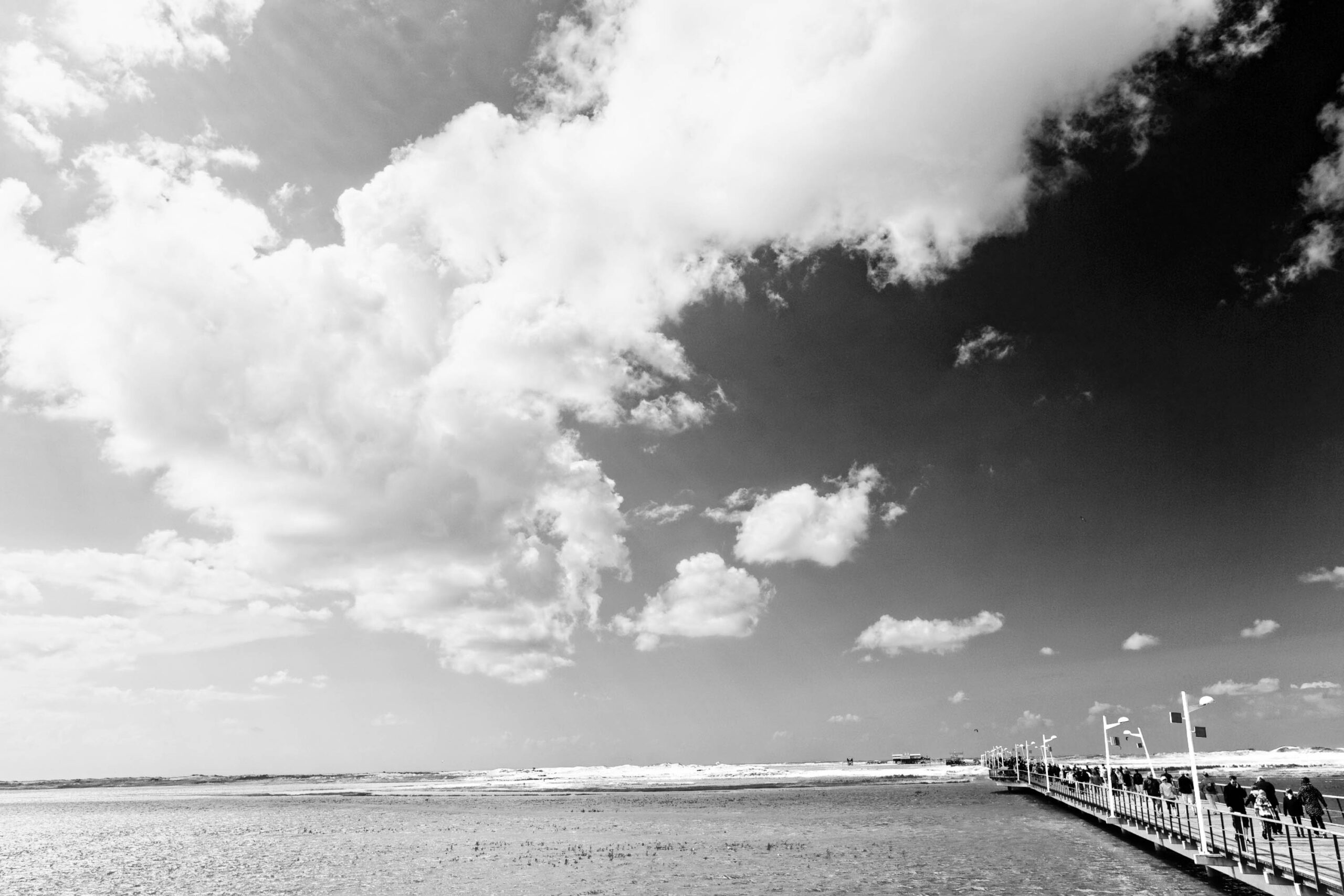 Das Foto zeigt eine Szene am Strand von St. Peter-Ording an der Nordsee. Ein hölzerner Steg erstreckt sich in den Himmel, der von dramatischen Wolkenformationen bedeckt ist. Das Wasser ist trüb und spiegelt das Licht wider. Im Hintergrund sind einige Personen auf dem Steg zu sehen. Die Szene vermittelt eine ruhige, aber auch etwas melancholische Stimmung.