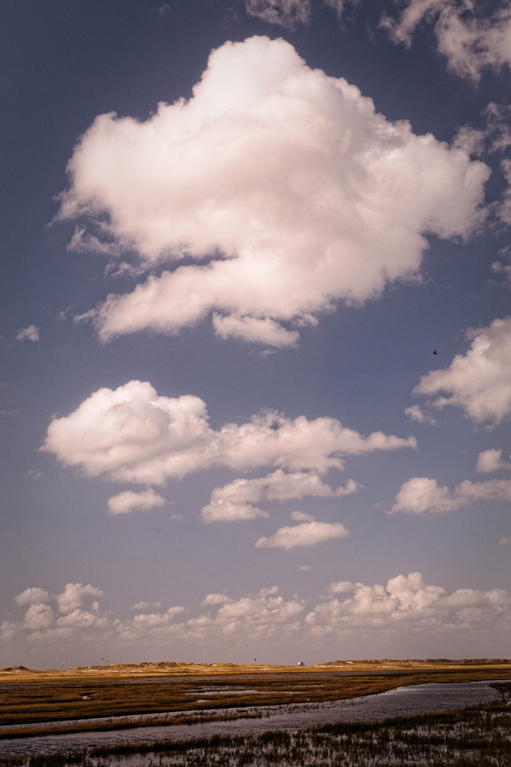 Die Aufnahme zeigt eine typische Landschaft der norddeutschen Küste, insbesondere von St. Peter-Ording. Im Vordergrund erstreckt sich eine weite, flache Gezeitengebiete, die bei Ebbe freiliegt. Das Wasser ist trüb und spiegelt das Licht der Wolken wider. Im Hintergrund ragen Dünen auf, die von einer spärlichen Vegetation bedeckt sind. Der Himmel ist von dramatischen Wolkenformationen dominiert, die das Licht reflektieren und einen starken Kontrast erzeugen. Die Szene vermittelt ein Gefühl von Weite, Ruhe und der Kraft der Natur.
