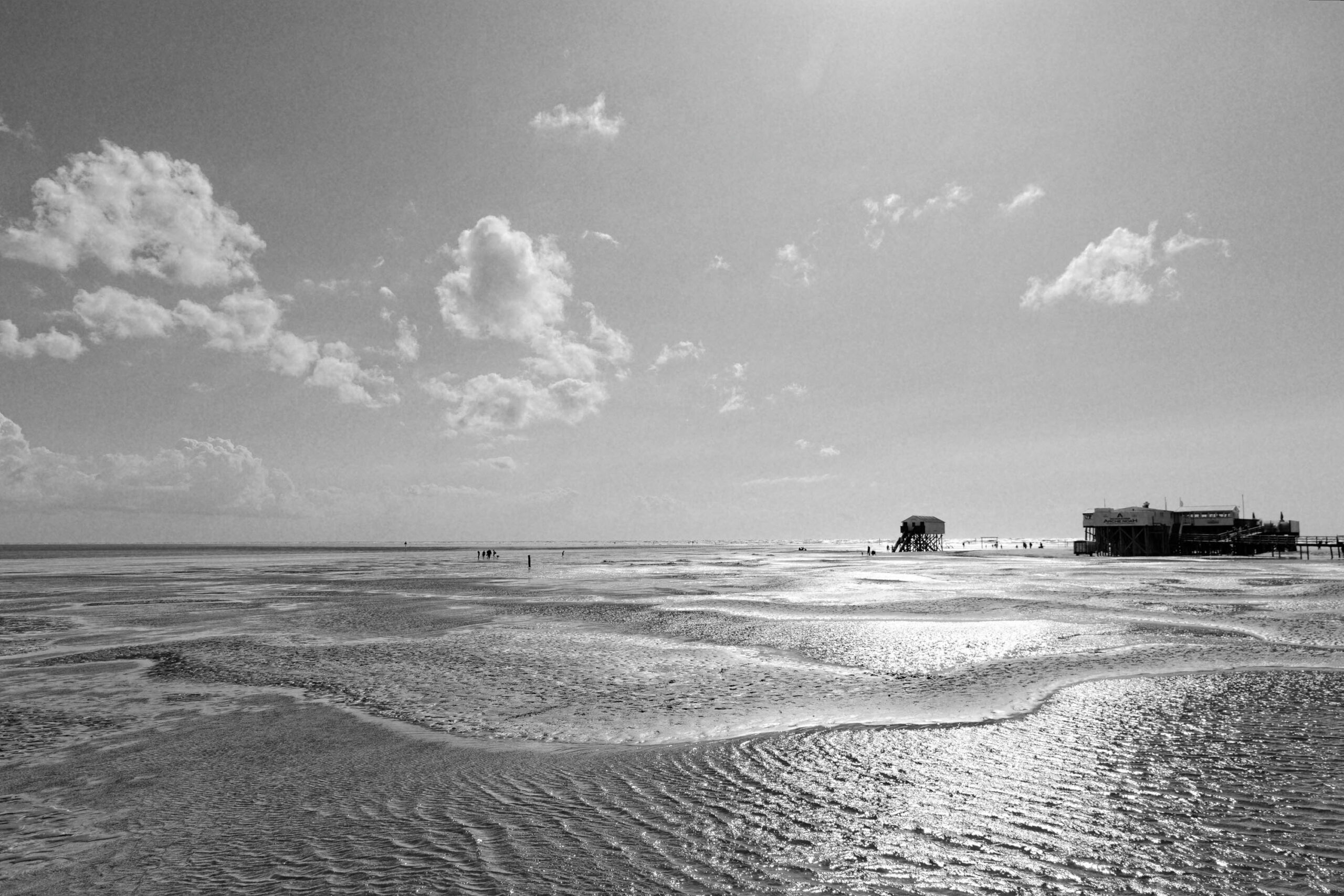Das Foto zeigt eine weite Strandlandschaft, wahrscheinlich bei Ebbe, mit glitzerndem Wasser und feuchtem Sand. Im Hintergrund sind mehrere Pfahlbauten (auch Strandkörbe genannt) zu sehen, die typisch für die Küste Norddeutschlands sind. Der Himmel ist bewölkt, aber die Sonne scheint durch die Wolken und erzeugt einen starken Kontrast. Die Szene vermittelt eine friedliche und entspannte Atmosphäre.