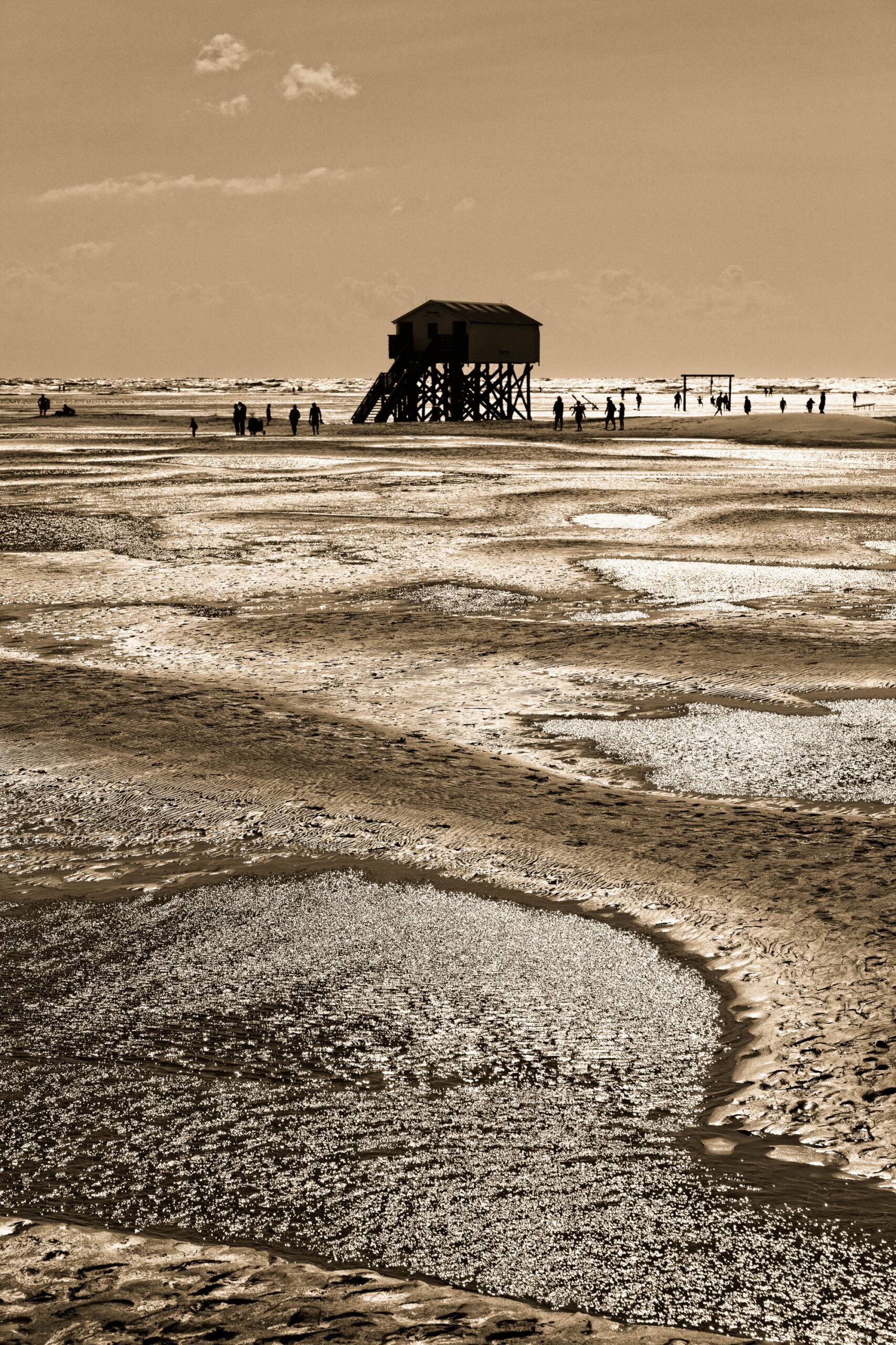Das vorliegende Schwarz-Weiß-Foto zeigt einen Pfahlbau auf dem weitläufigen Strand von St. Peter-Ording. Der Pfahlbau, ein charakteristisches Merkmal der Küste, steht auf langen Holzpfählen und bietet einen erhöhten Aussichtspunkt. Der Strand erstreckt sich bis zum Horizont, durchzogen von Wellen, die interessante Muster im Sand hinterlassen. Eine Gruppe von Menschen befindet sich in der Nähe des Pfahlbaus und scheint die Landschaft zu genießen. Das Licht ist weich und erzeugt eine ruhige und friedliche Atmosphäre. Die Komposition des Fotos betont die Weite des Strandes und die Isolation des Pfahlbaus.