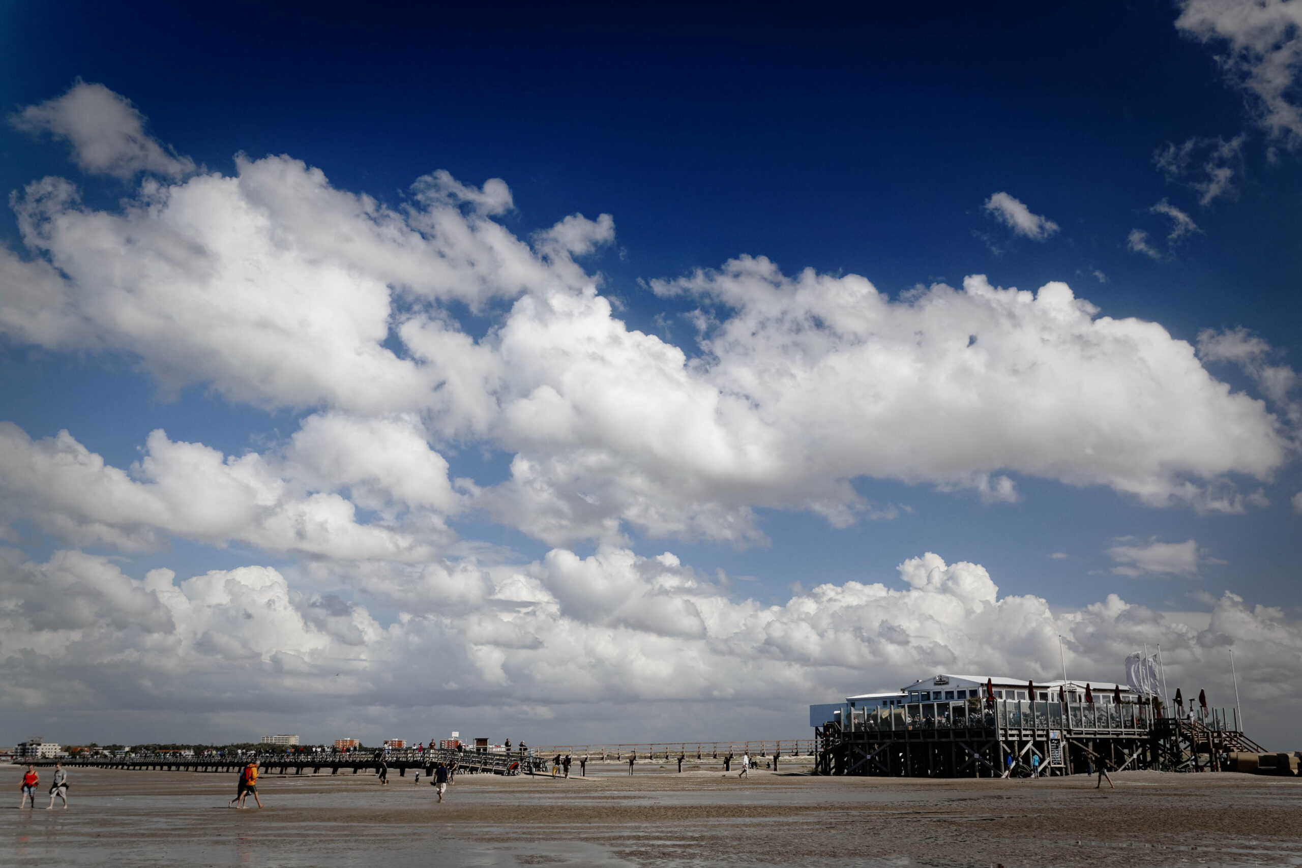 Die Aufnahme zeigt eine weitläufige Szene am Strand von St. Peter-Ording, Deutschland. Im Vordergrund erstreckt sich ein breiter Sandstrand, der von wenigen Personen besucht wird. Im Hintergrund erhebt sich ein markanter Pfahlbau, der typisch für diese Küstenregion ist. Der Himmel ist von dichten, weißen Wolken bedeckt, die einen dynamischen Kontrast zum dunklen Untergrund bilden. Die Perspektive ist weitwinkelig, was die Weite des Strandes und des Himmels betont. Die Beleuchtung ist diffus, was eine sanfte Atmosphäre schafft.