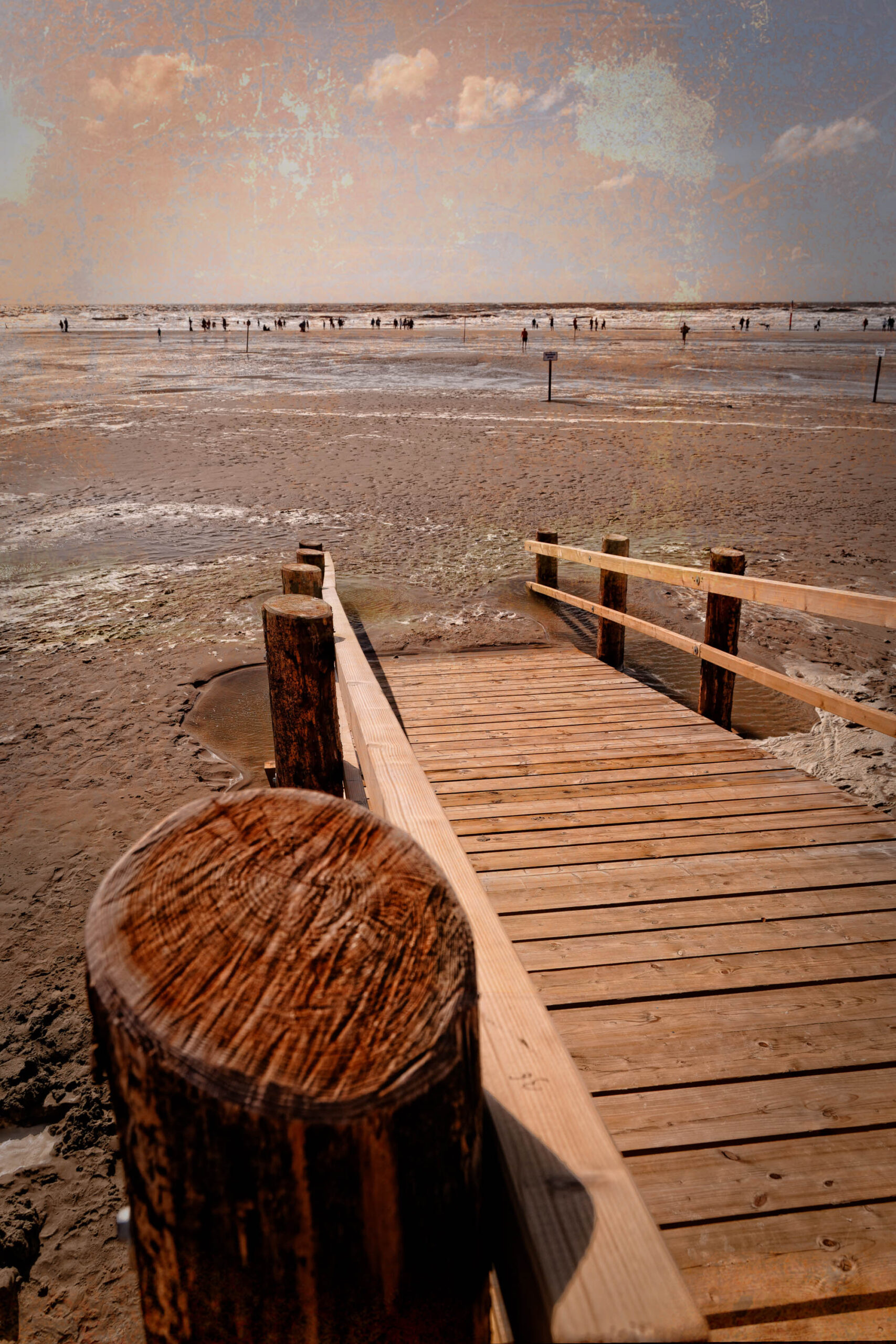 Das Bild zeigt einen hölzernen Steg, der sich in die Ferne eines Sandstrandes erstreckt. Der Steg ist aus Holz und hat eine einfache Konstruktion mit gelagerten Holzbalken als Geländer. Der Strand ist weitläufig und sandig, mit einigen Personen im Hintergrund, die spazieren gehen. Der Himmel ist bewölkt und der Sonnenuntergang verleiht der Szene eine warme, goldene Beleuchtung. Die Szene vermittelt ein Gefühl von Ruhe und Weite.
