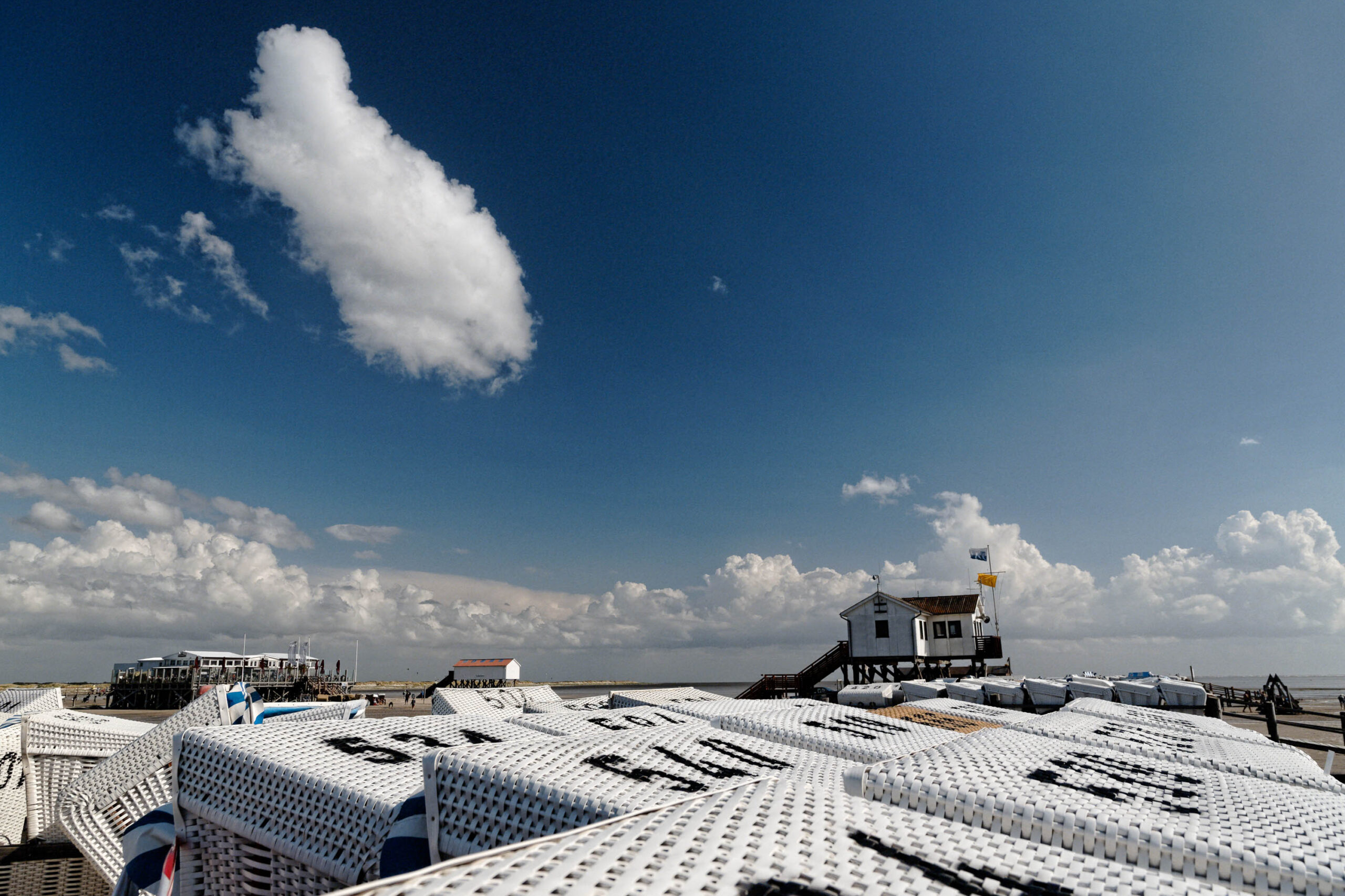 Die Aufnahme zeigt eine malerische Szene am Strand von St. Peter-Ording, Deutschland. Im Vordergrund dominieren die charakteristischen weißen Pfahlbauten, die typisch für diese Region sind. Die Pfahlbauten sind dicht aneinander gereiht und bilden ein interessantes Muster. Im Hintergrund erstreckt sich ein strahlend blauer Himmel mit vereinzelten weißen Wolken. Die Szene vermittelt eine sommerliche und entspannte Atmosphäre.