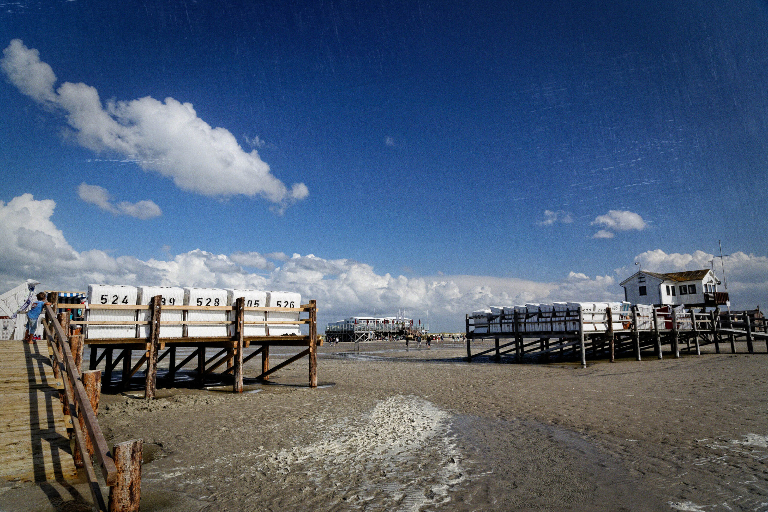 Die Aufnahme zeigt eine Reihe von traditionellen Strandkörben, die auf dem Sandstrand von St. Peter-Ording, Nordsee, aufgestellt sind. Die Strandkörbe sind in einer Reihe angeordnet und stehen auf Holzpfählen. Der Himmel ist blau und von weißen Wolken bedeckt. Im Hintergrund sind weitere Strandkörbe und Gebäude zu sehen. Der Strand ist feucht und glänzend, was auf Ebbe hindeutet. Die Szene vermittelt eine friedliche und entspannte Atmosphäre.