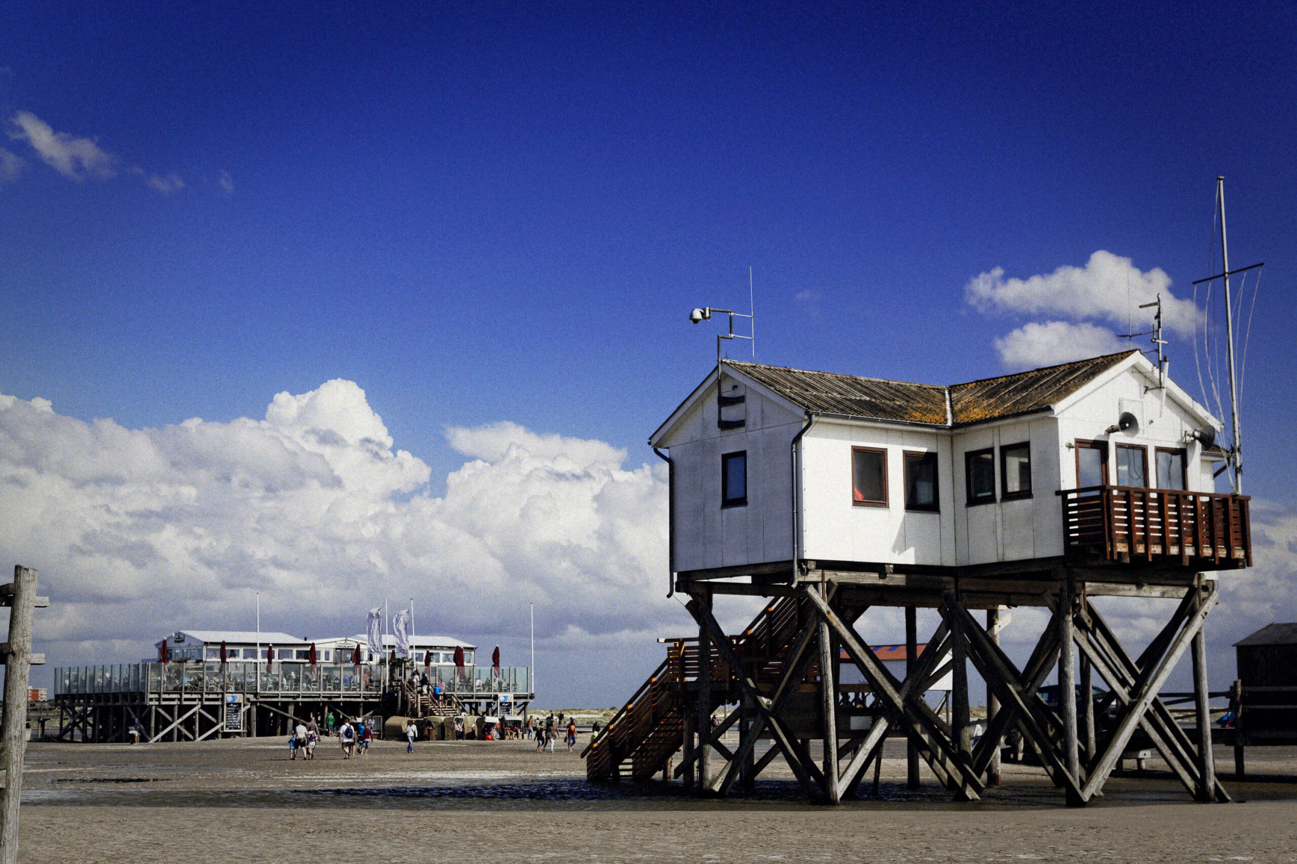Das Bild zeigt einen typischen Pfahlbau am Strand von St. Peter-Ording. Der Bau ist weiß gestrichen und steht auf stabilen Holzpfählen. Im Hintergrund sind weitere Pfahlbauten sowie ein Strand mit Menschen erkennbar. Der Himmel ist blau mit vereinzelten weißen Wolken. Der Strand ist sandig und weitläufig.
