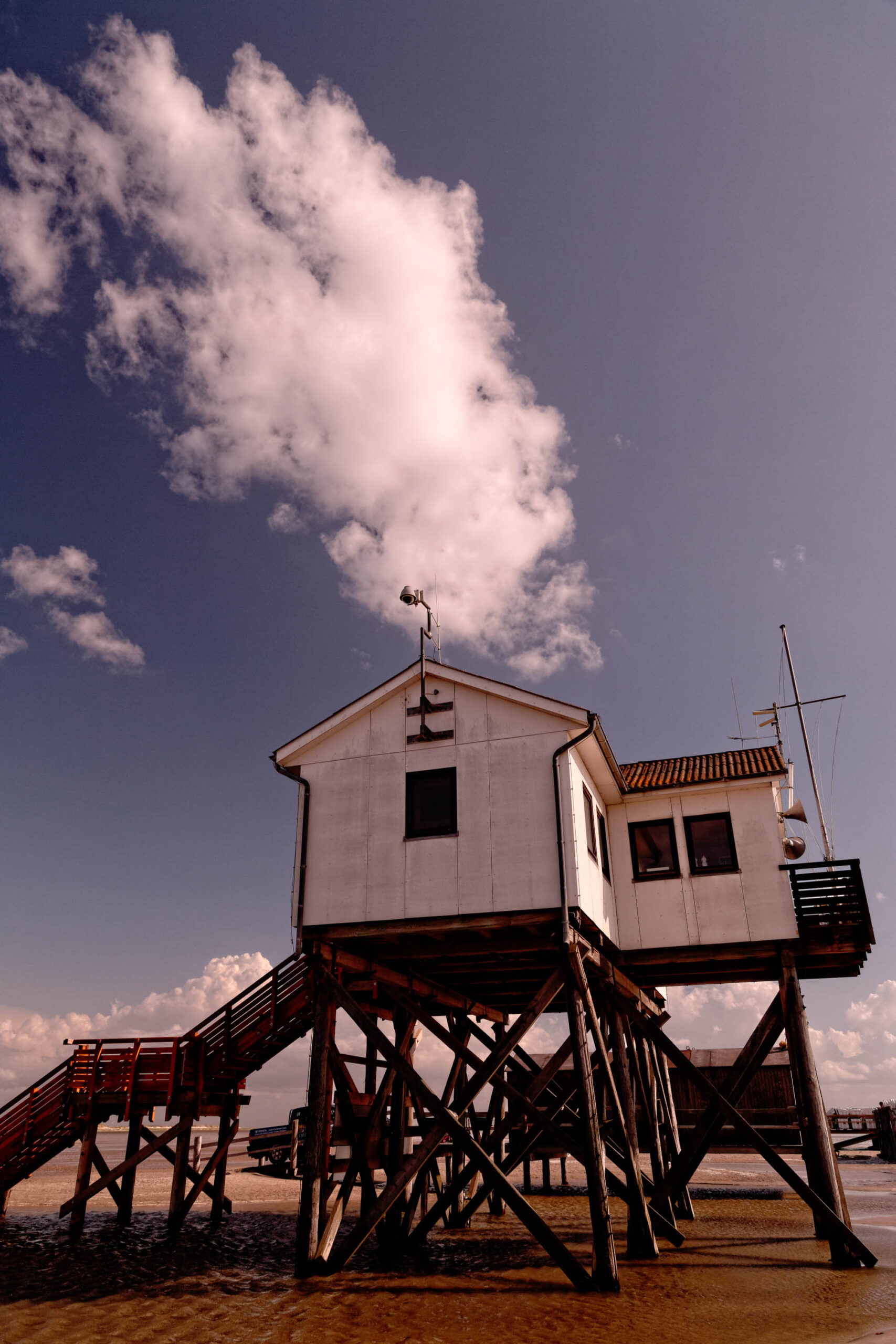 Das Bild zeigt einen typischen Pfahlbau am Strand von St. Peter-Ording. Der Bau ist weiß gestrichen und steht auf dunklen Holzpfählen, die ihn deutlich über den Sandstrand erheben. Die Konstruktion ist mit einer Treppe erreichbar. Der Himmel ist von dicken, weißen Wolken bedeckt, was eine dramatische Lichtstimmung erzeugt. Der Strand ist sandig und leicht feucht. Die Perspektive ist von unten, was die Höhe des Pfahlbaus betont. Es handelt sich um eine typische Szene des Ordinger Strandes, die für ihre charakteristischen Pfahlbauten bekannt ist.