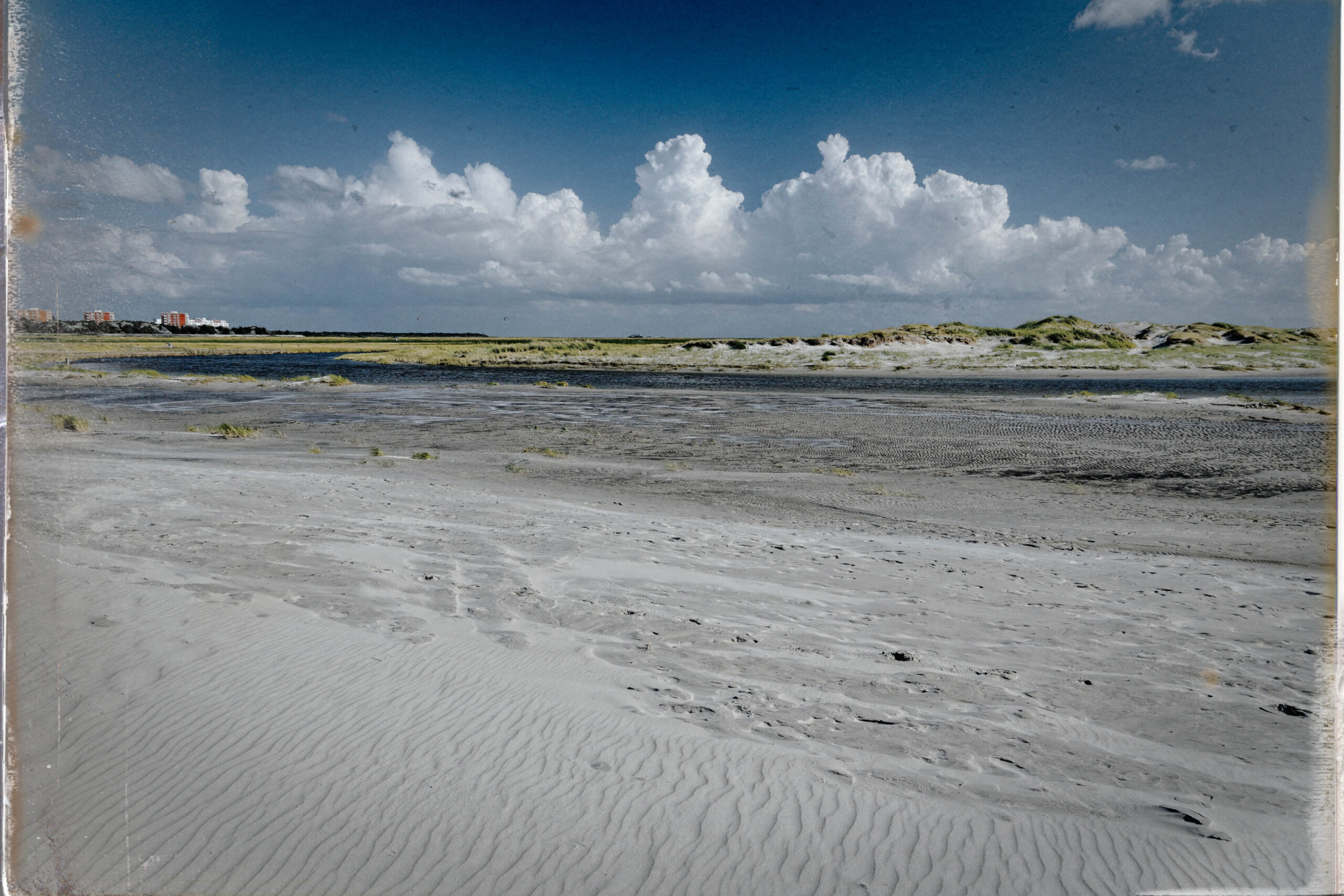 Die Aufnahme zeigt eine weite Sandfläche, die von Wind geformten Wellenmustern. Im Hintergrund erheben sich Dünen, die mit spärlicher Vegetation bedeckt sind. Der Himmel ist blau und von weißen Wolken bedeckt. Die Szene vermittelt ein Gefühl von Weite und Ruhe.