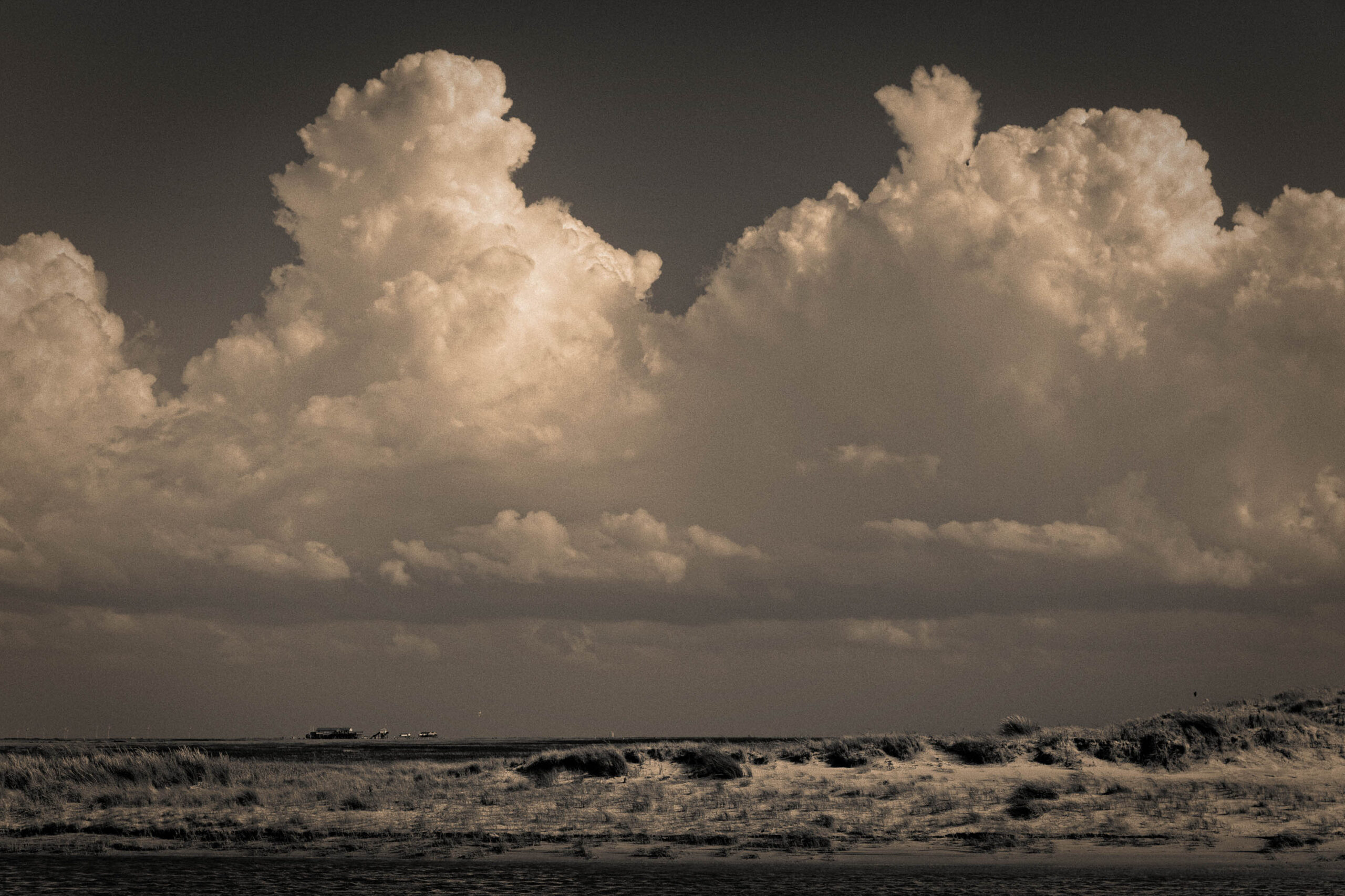 Das Foto zeigt eine dramatische Szene an einem norddeutschen Strand, wahrscheinlich in St. Peter-Ording. Der Himmel ist von dicken, weißen Wolken bedeckt, die einen starken Kontrast zum dunklen Wasser und dem dunklen Sand bilden. Die Wellen brechen auf dem Strand und erzeugen eine dynamische Atmosphäre. Im Hintergrund sind mehrere Windkraftanlagen zu sehen, die die industrielle Präsenz in der Landschaft unterstreichen. Die monochrome Darstellung verstärkt die Stimmung von Kraft und Weite.
