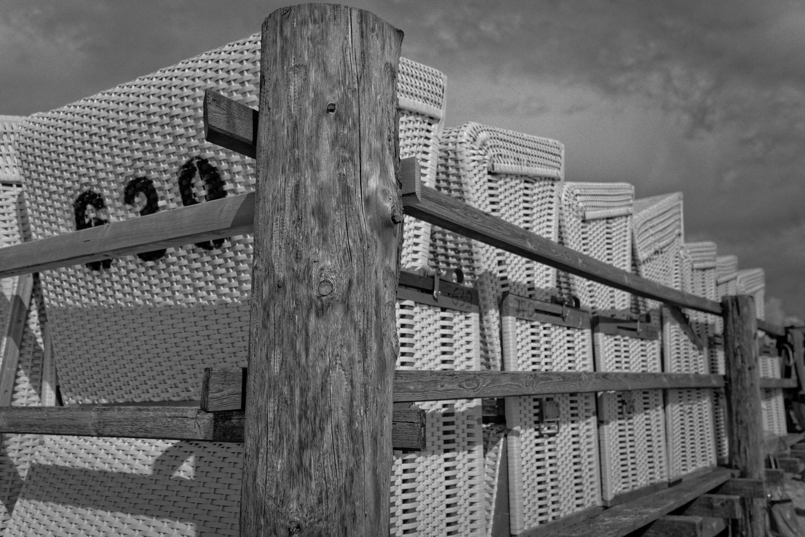 Das Foto zeigt einen beeindruckenden Anblick von Strohdächern, die in einer terrassenartigen Anordnung nach oben führen. Die Dächer sind dicht aneinander gereiht und erzeugen ein faszinierendes Muster. Im Vordergrund befindet sich ein grob gezimmertes Holzgeländer mit einem dicken Holzpfosten, das die Szene rahmt. Das Licht ist diffus und erzeugt einen starken Kontrast zwischen den hellen Strohdächern und den dunklen Schatten. Die Komposition ist interessant, da sie die vertikale Anordnung der Dächer betont und gleichzeitig die Textur des Holzes hervorhebt. Das Bild vermittelt ein Gefühl von Ruhe und Beständigkeit, aber auch von der rauen Schönheit der norddeutschen Küste.