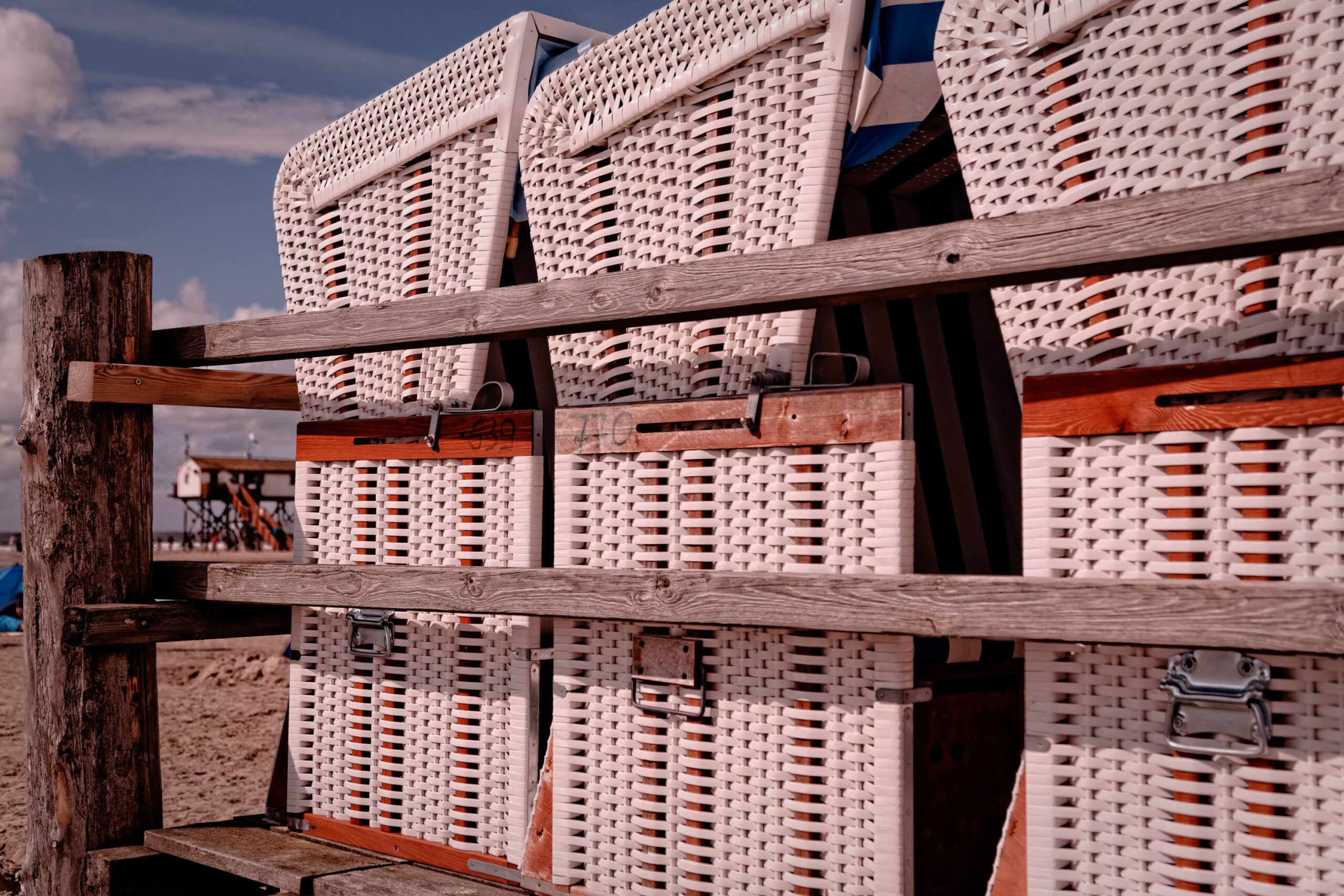 Das Bild zeigt eine Reihe von traditionellen Korbtassen, die auf einer Holzkonstruktion gestapelt sind. Die Korbtassen sind weiß und haben braune Griffe. Sie sind typisch für die Region St. Peter-Ording an der Nordsee. Im Hintergrund ist der Strand und der Himmel zu sehen. Das Bild vermittelt eine ruhige und entspannte Atmosphäre.