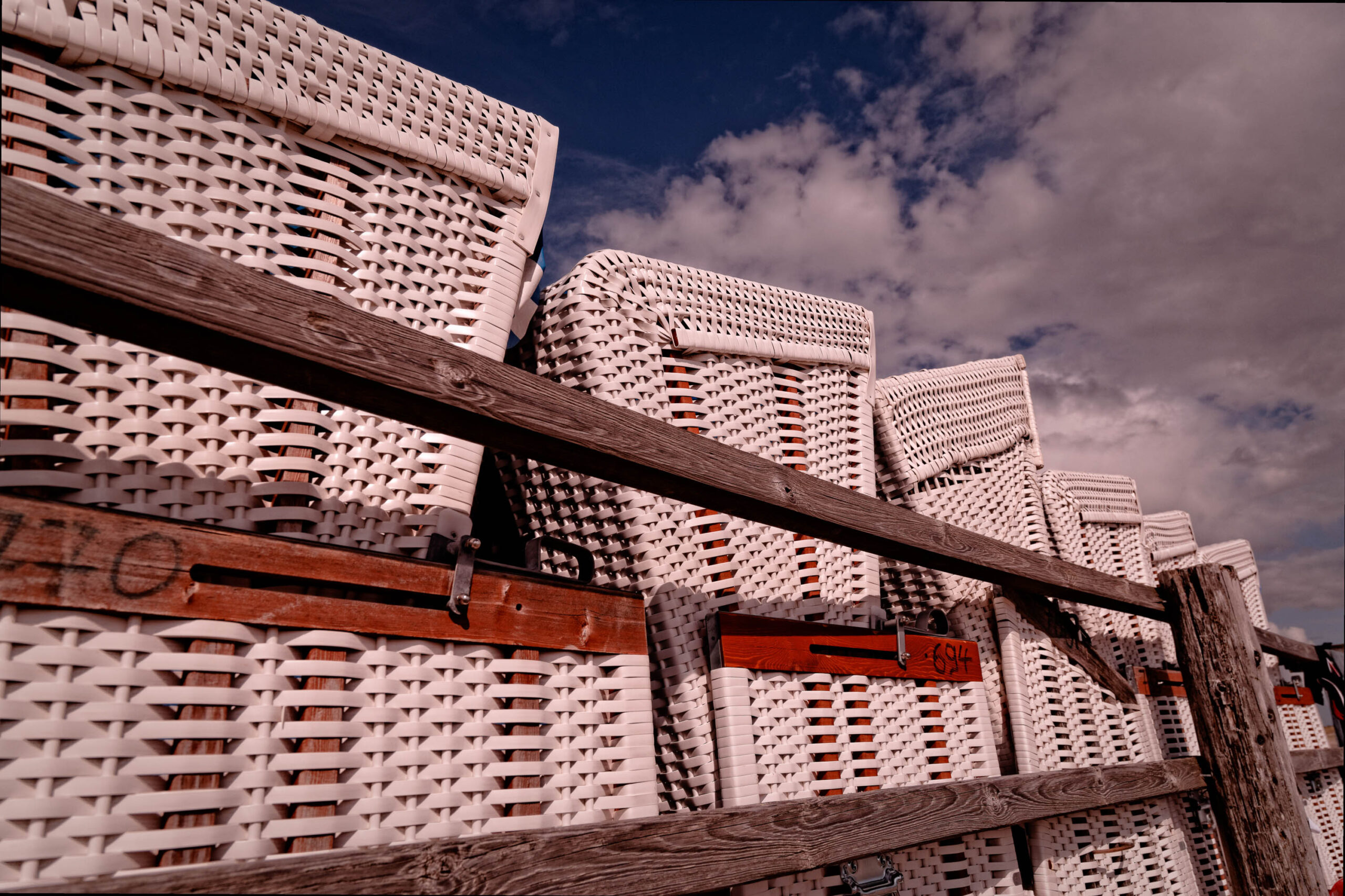 Das Bild zeigt eine ungewöhnliche Perspektive auf gestapelte Kunststoffkörbe, die sich in einer Art Pyramide aufgeschichtet sind. Die Körbe sind weiß und haben ein charakteristisches, gitterartiges Muster. Sie stehen vor einem dunklen, bewölkten Himmel, der einen starken Kontrast zum hellen Weiß der Körbe bildet. Ein hölzerner Zaun verläuft diagonal durch das Bild und rahmen die Körbe ein. Die Körbe scheinen in St. Peter-Ording, Nordsee, zu stehen, was durch die typische Landschaft und die Art der Körbe bestätigt wird. Die Körbe sind wahrscheinlich für den Transport von Strandgut oder für andere landwirtschaftliche Zwecke gedacht.