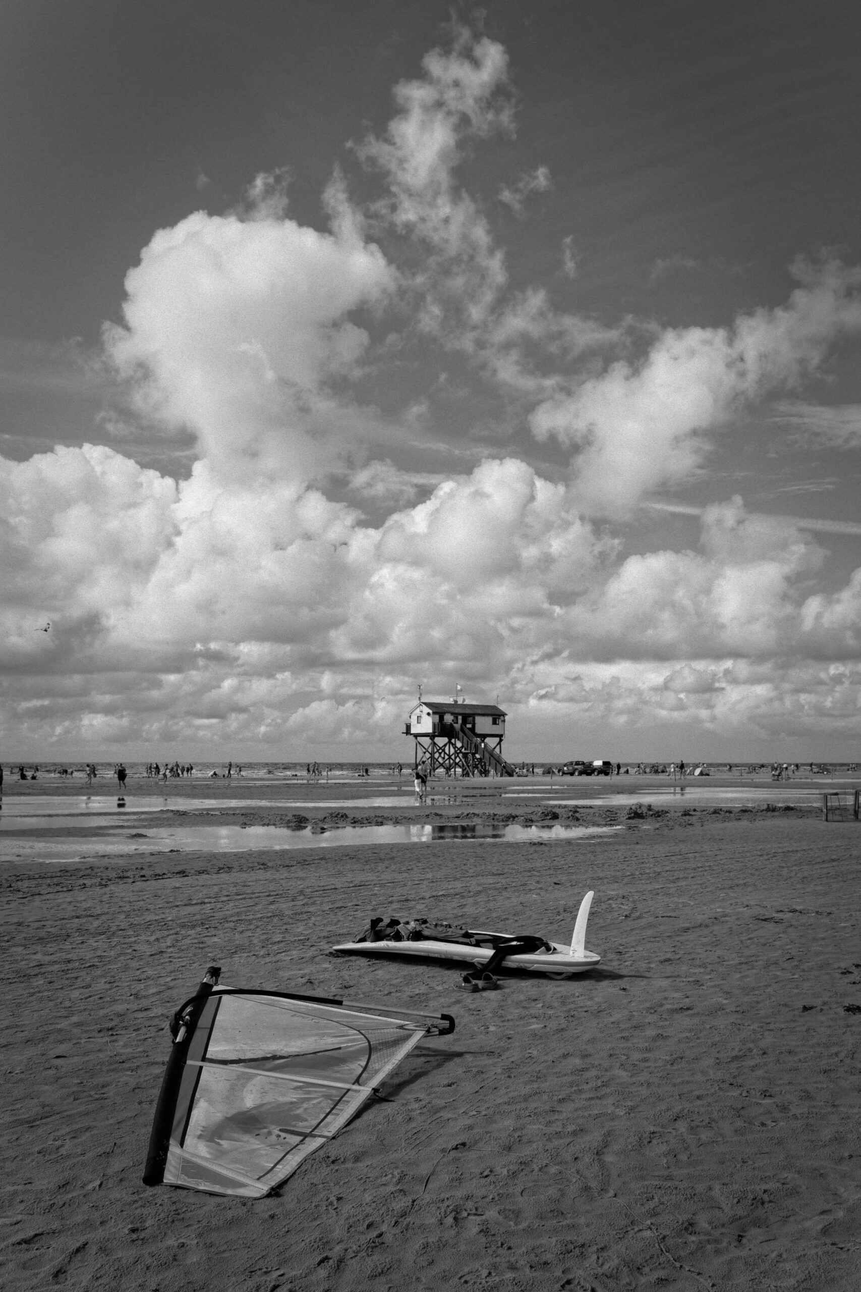 Das Foto zeigt eine Szene am Strand von St. Peter-Ording, Deutschland. Im Vordergrund stehen mehrere Windsurfbretter, die teilweise im Sand liegen. Im Hintergrund ist ein erhöhter Strandwachtturm (Pfahlbau) zu sehen, der auf Stelzen steht. Der Himmel ist bewölkt und erzeugt einen dramatischen Kontrast zum hellen Sandstrand. Menschen sind im Hintergrund zu erkennen, die sich am Strand aufhalten.