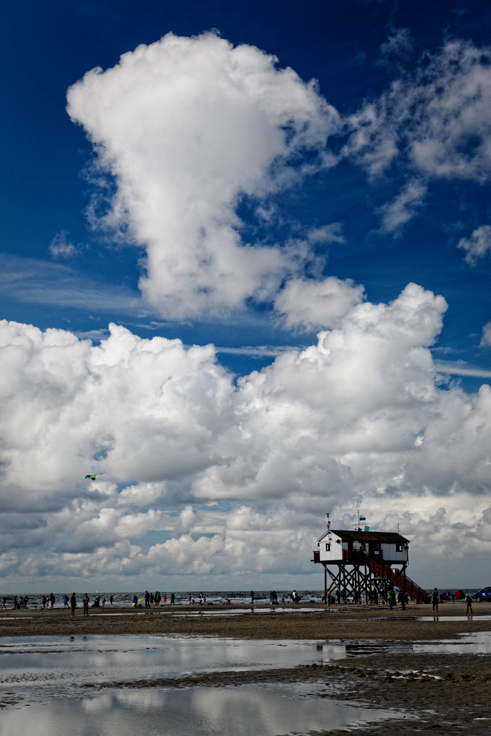 Das Foto zeigt einen Pfahlbau am Strand von St. Peter-Ording. Der Himmel ist bewölkt und der Strand ist teilweise mit Wasser bedeckt, was eine Spiegelung des Himmels erzeugt. Im Hintergrund sind Personen zu sehen, die sich auf dem Strand aufhalten. Die Szene vermittelt eine ruhige und entspannte Atmosphäre.