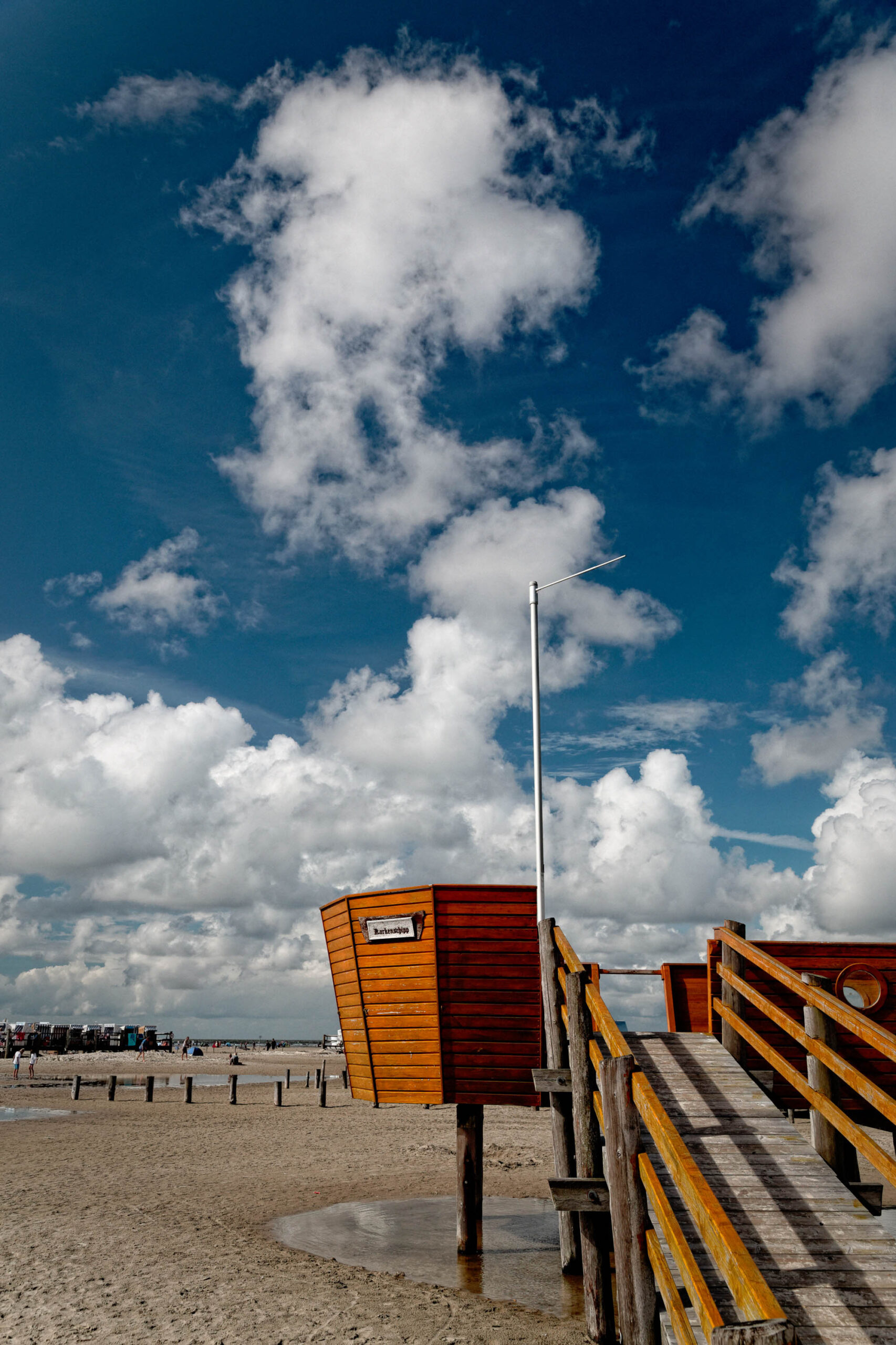 Das Bild zeigt einen typischen Pfahlbau (ein Gebäude auf Stelzen) am Strand von St. Peter-Ording. Der Bau ist aus Holz und hat ein Schild mit der Aufschrift „Kulturdepot“. Im Hintergrund erstreckt sich ein breiter Sandstrand, der von einer Reihe von Personen und Sonnenschirmen besetzt ist. Der Himmel ist dramatisch mit dicken, weißen Wolken vor einem tiefblauen Hintergrund. Die Perspektive ist von unten, was die Höhe des Pfahlbaus und die Weite des Himmels betont.