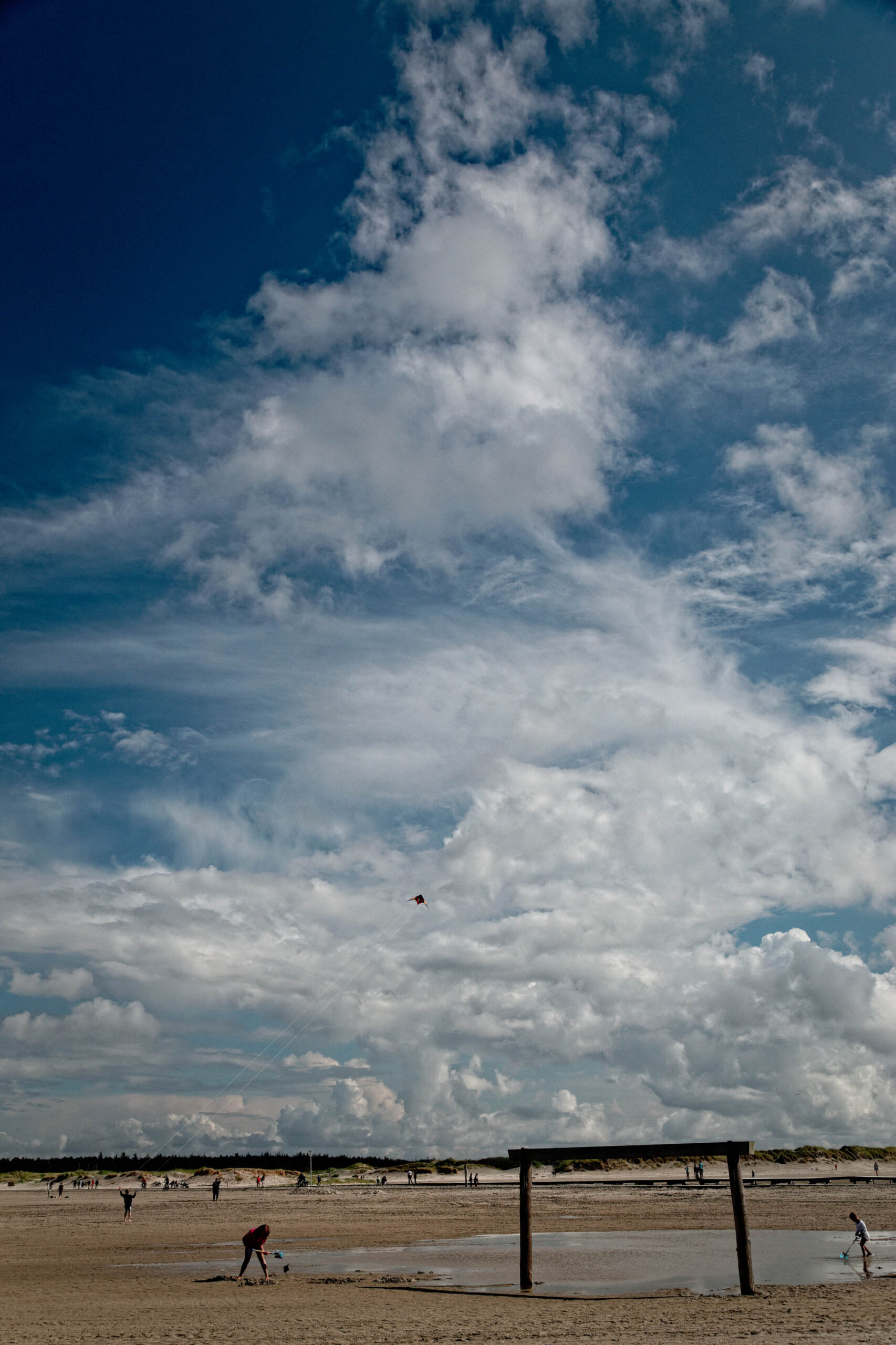 Die Aufnahme zeigt eine typische Szene von St. Peter-Ording an der Nordsee. Der weite Sandstrand erstreckt sich bis zum Horizont, wo er in den Himmel übergeht, der von zahlreichen, weißen Wolken bedeckt ist. Im Vordergrund sind Personen zu sehen, die sich auf dem Strand aufhalten und verschiedene Aktivitäten ausüben. Im Hintergrund sind Dünen zu erkennen, die die Landschaft prägen. Die Aufnahme vermittelt ein Gefühl von Weite und Freiheit.