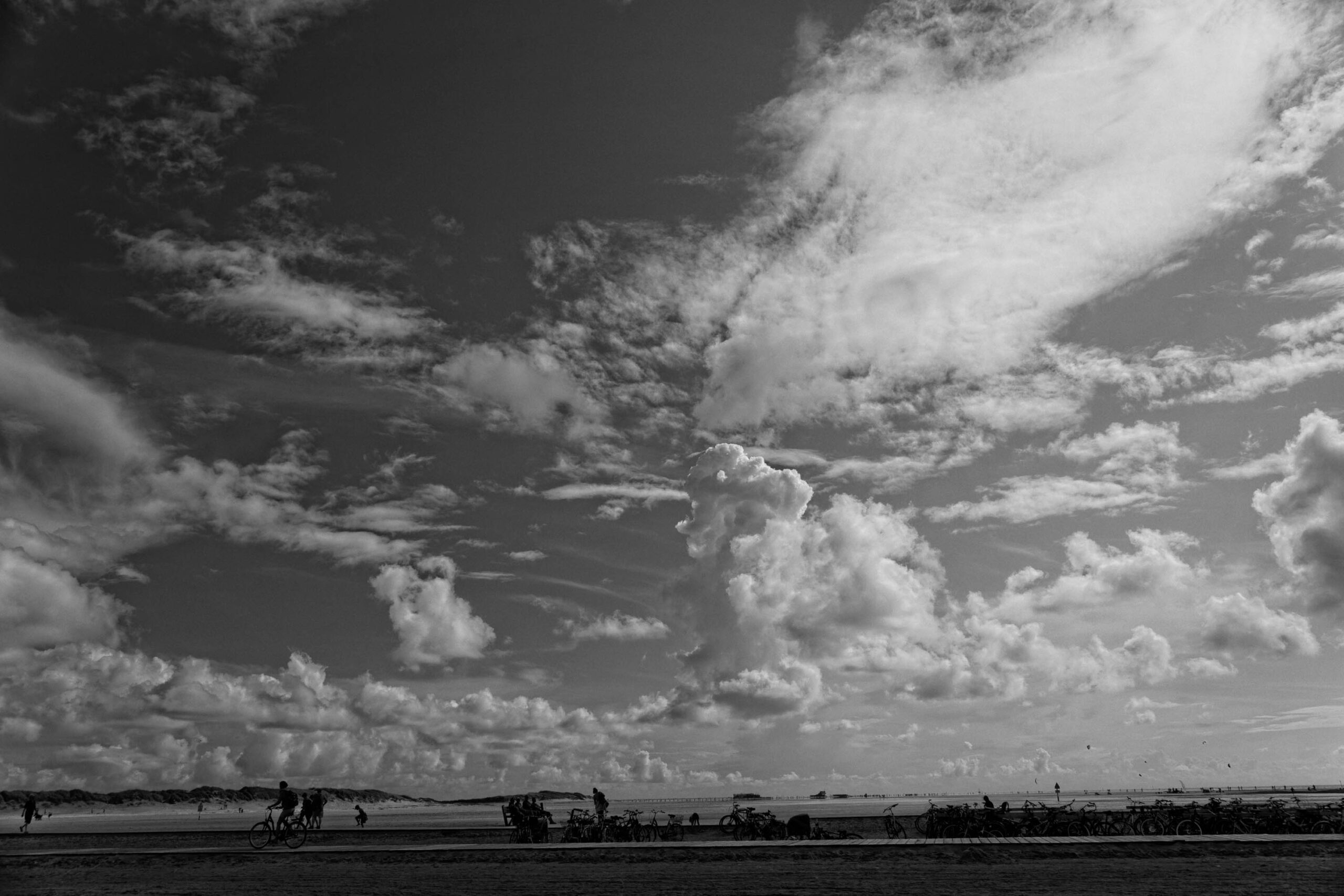 Die Fotografie fängt eine Szene am Strand von St. Peter-Ording ein, wo eine große Anzahl von Fahrrädern und Menschen versammelt ist. Der Himmel ist von einer beeindruckenden Wolkenformation bedeckt, die einen starken Kontrast zum dunklen Strand erzeugt. Die Szene vermittelt eine Atmosphäre von Erholung und Aktivität, während die Schwarz-Weiß-Darstellung einen nostalgischen und dramatischen Effekt erzeugt.