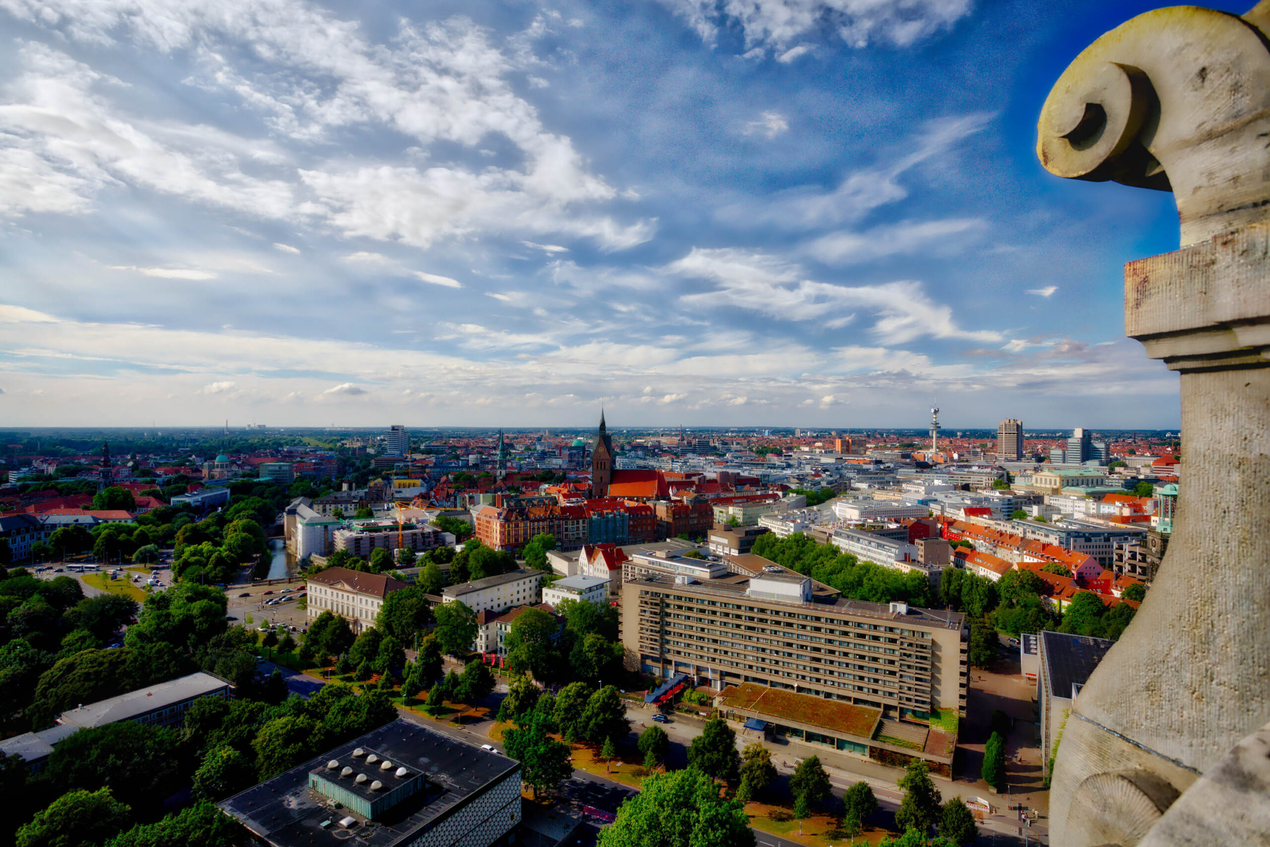 Die Aufnahme zeigt eine weitläufige Stadtlandschaft, die sich bis zum Horizont erstreckt. Die Stadt ist von grünen Bäumen und Parks umgeben. Im Vordergrund befindet sich ein Gebäude mit einer steinernen Fassade. Im Hintergrund sind mehrere Gebäude unterschiedlicher Höhe und Architektur zu sehen. Der Himmel ist bewölkt, aber die Sonne scheint durch die Wolken und beleuchtet die Stadt. Die Aufnahme ist in einem warmen Licht gehalten, das die Stadt in ein positives Licht rückt.